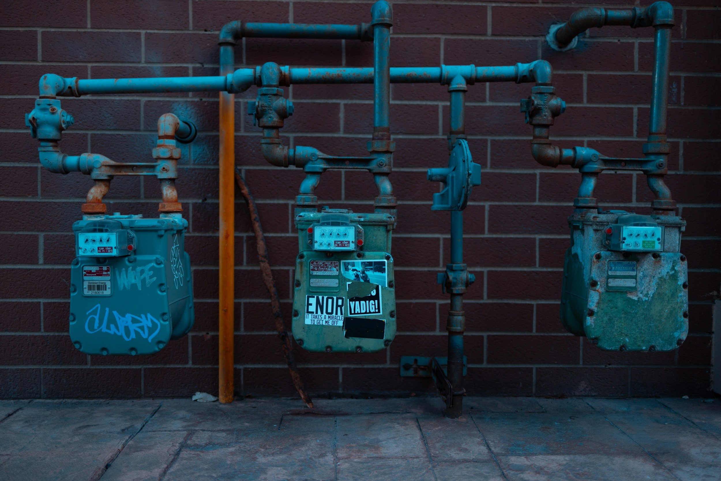 Three utility meters attached to a brick wall, with pipes running above them. Two meters are blue, and one is gray with stickers and graffiti. The meters have digital displays and are secured with locks and stickers, with some graffiti and stickers on them.