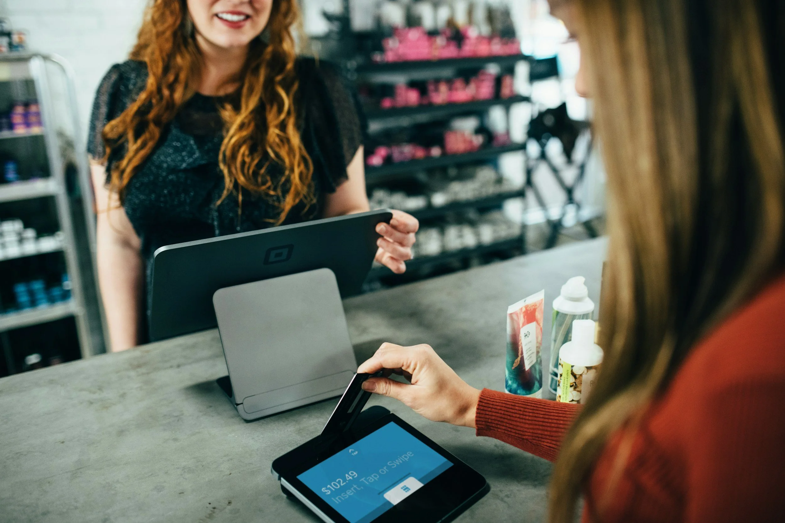A store clerk smiling and handing a credit card reader to a customer, who is holding a card reader device, at a checkout counter.