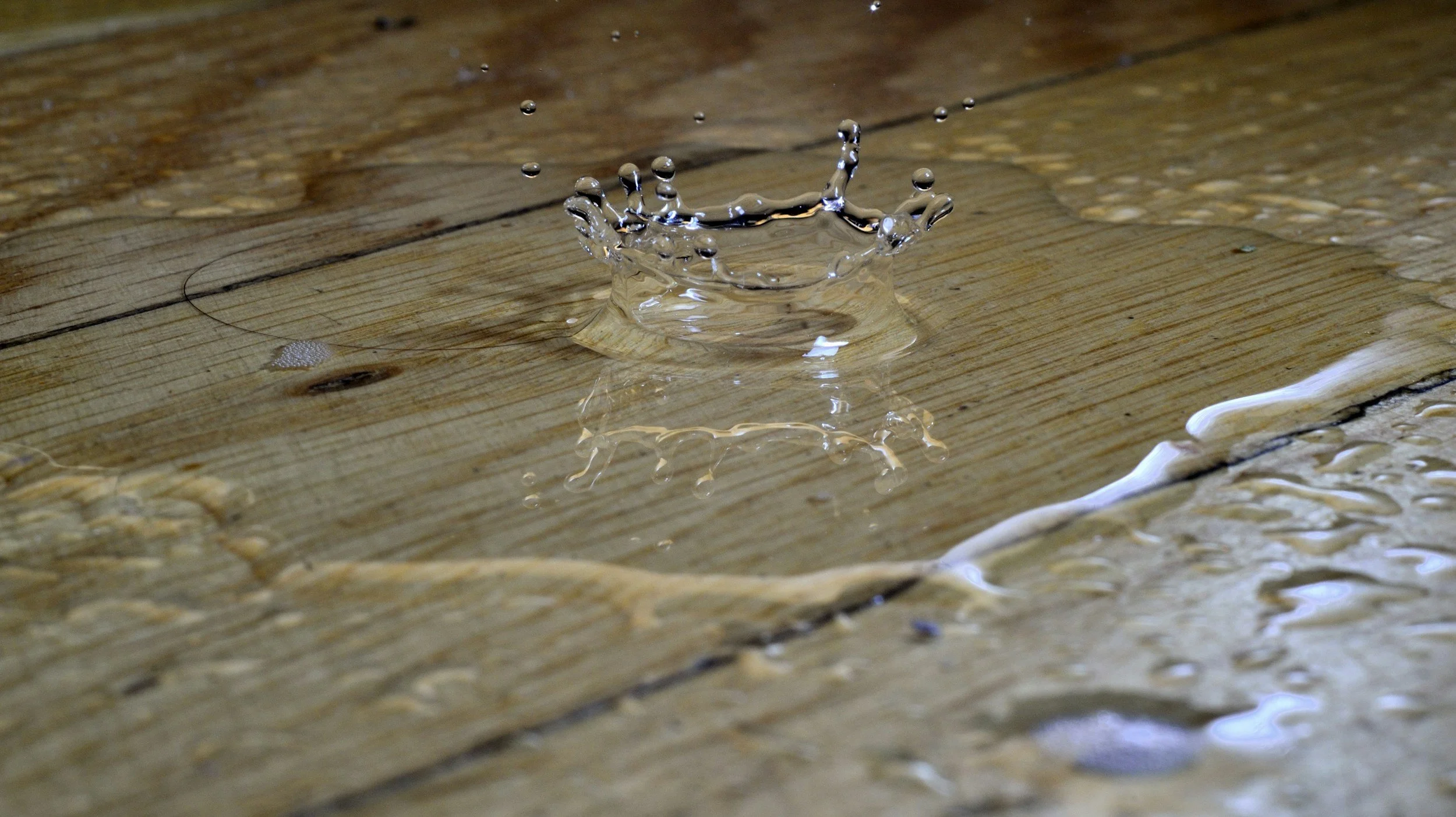 A close-up of a water droplet splashing on a wooden surface, forming a crown-shaped splash and reflected on the surface below.