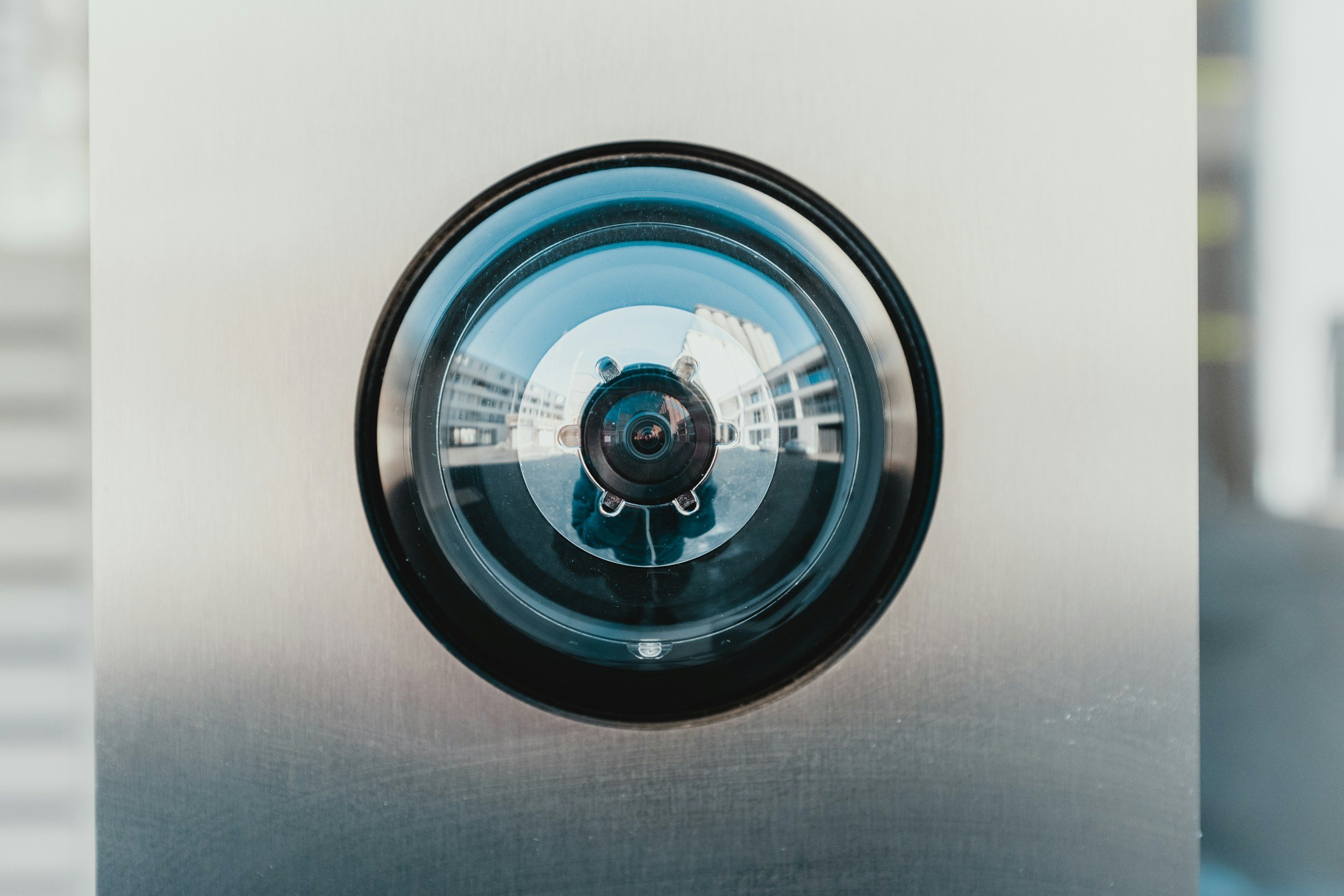 Close-up of a security camera mounted on a building exterior, reflecting surrounding buildings and sky.