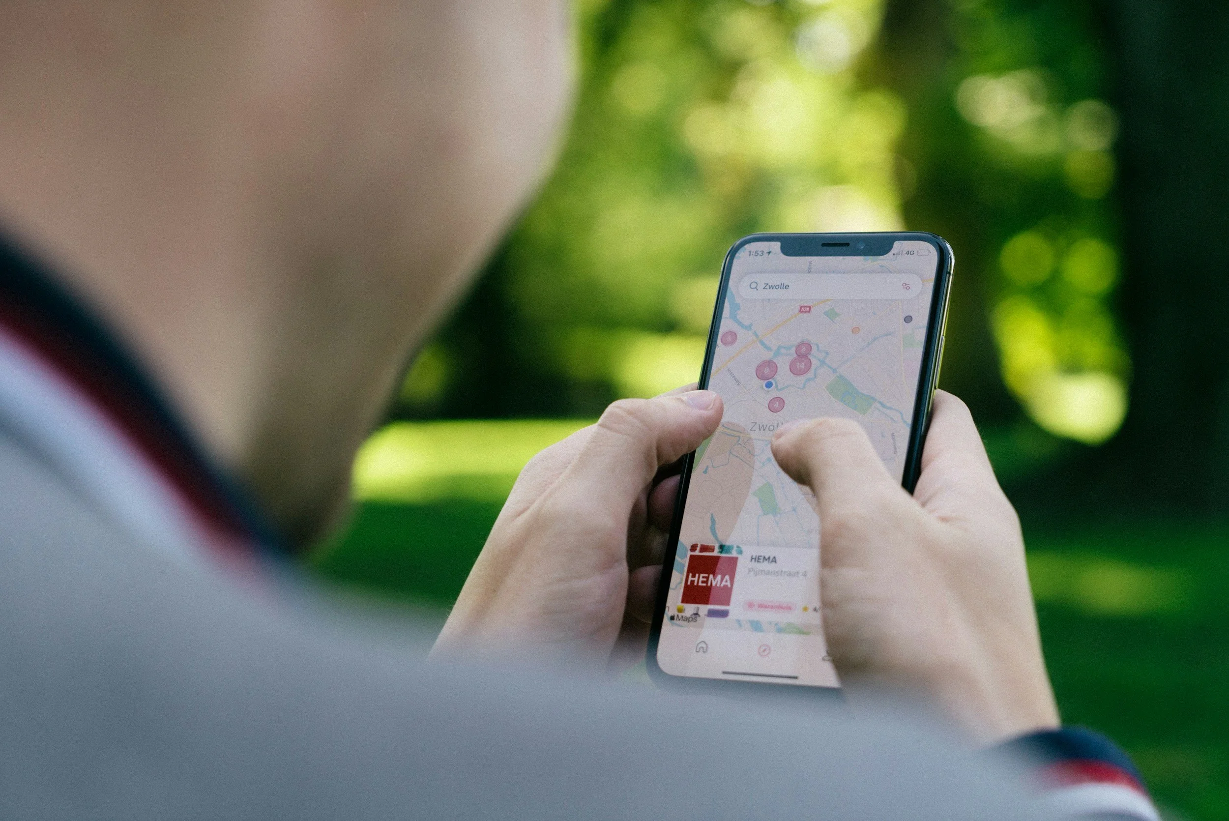 Person holding a smartphone showing a map with multiple location markers, outdoors with greenery in the background.