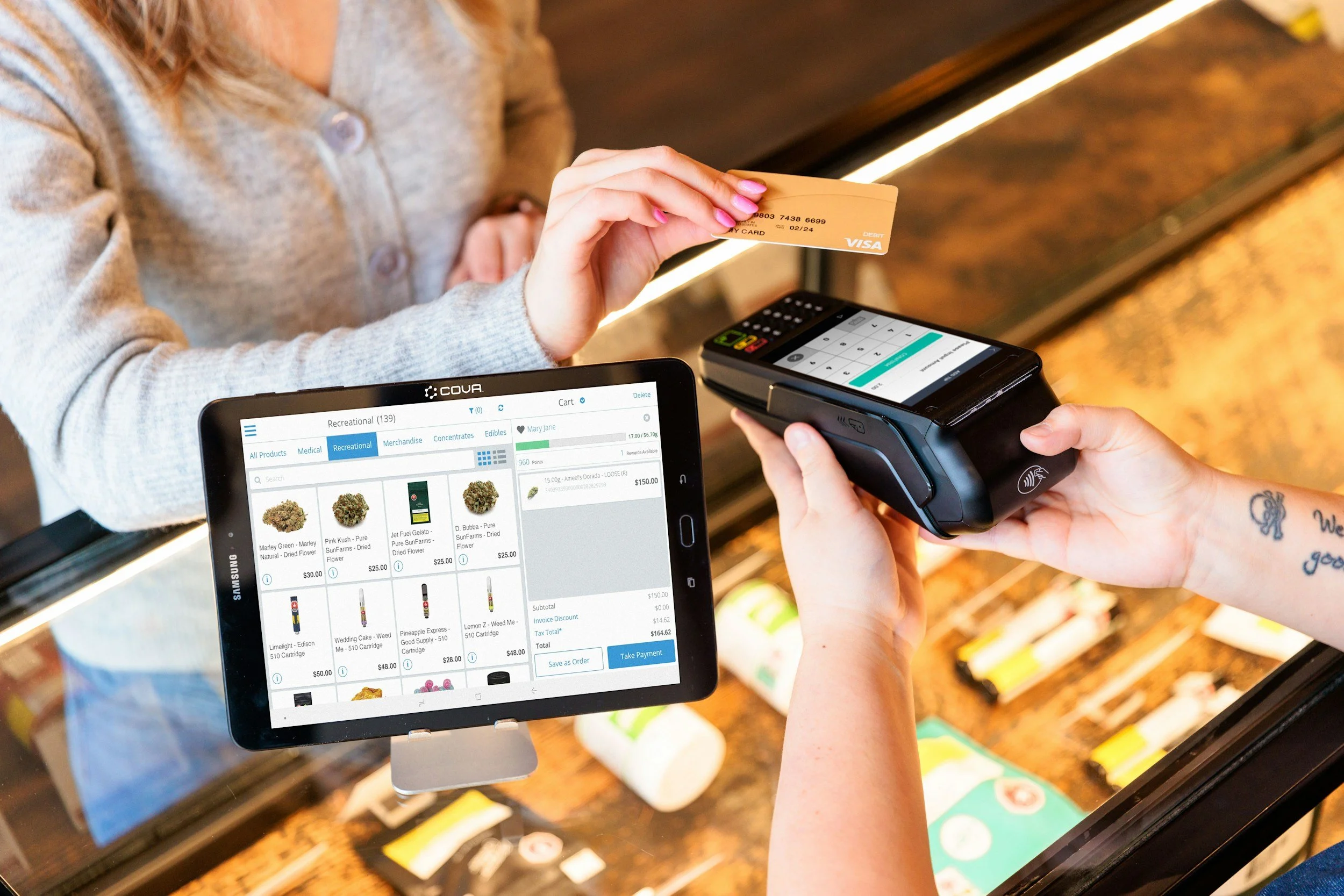 Person holding a yellow credit card at the checkout counter while a cashier processes a payment with a point-of-sale terminal, and a tablet displaying an online shopping cart with marijuana products is visible in the foreground.