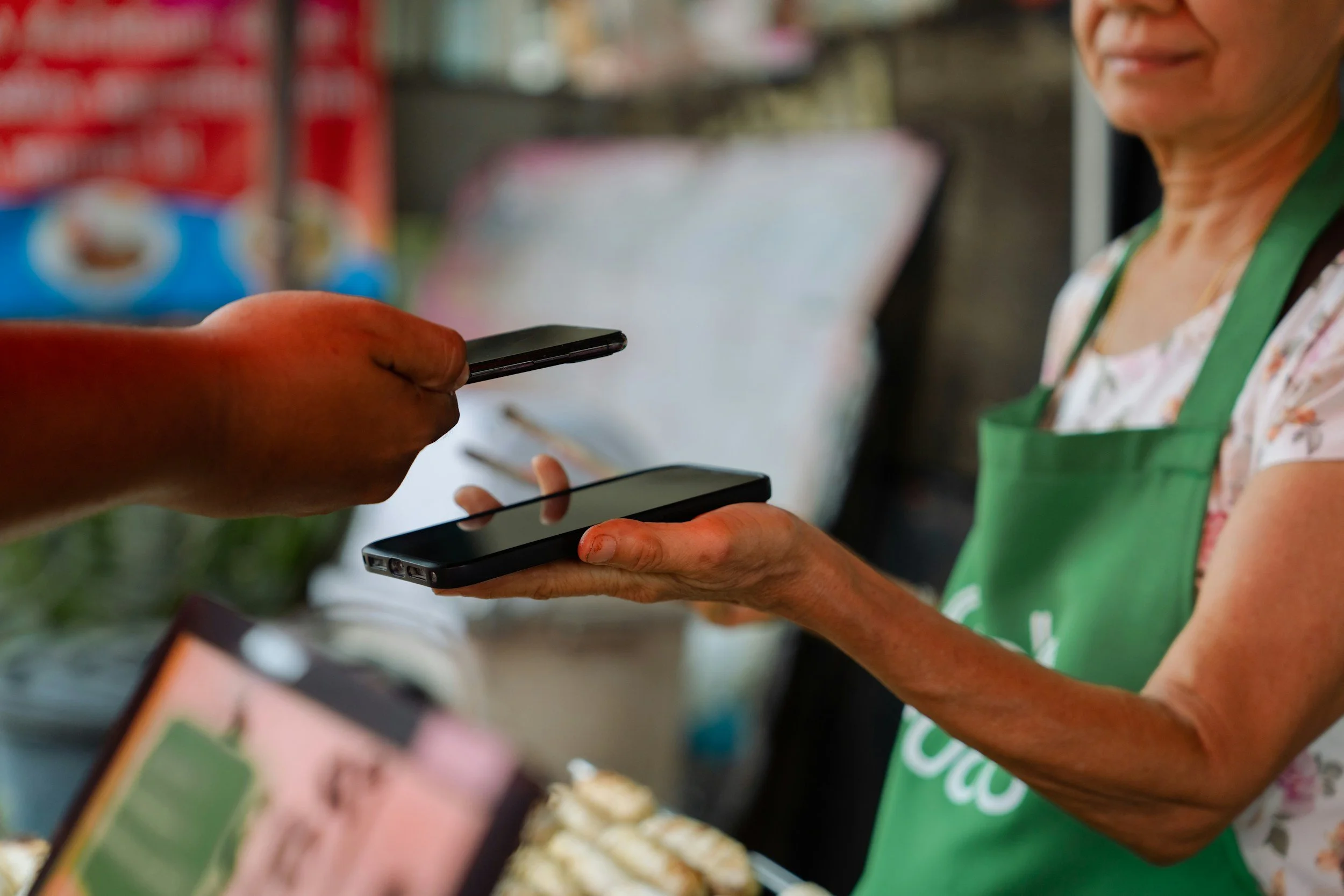 A person in a green apron is holding a smartphone, with another person handing them their phone in a busy outdoor setting with snacks or food displayed in the background.