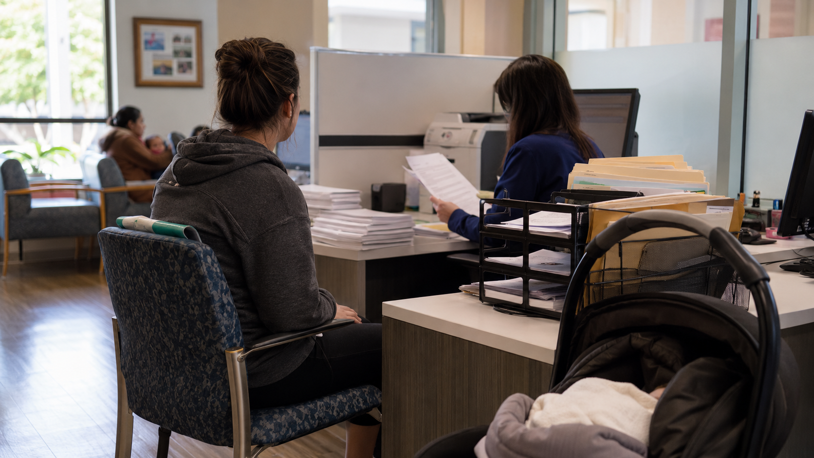 A new mother waits with an infant stroller while administrative staff review paperwork at a public services office, illustrating the legal and child welfare process that can follow postpartum THC testing.