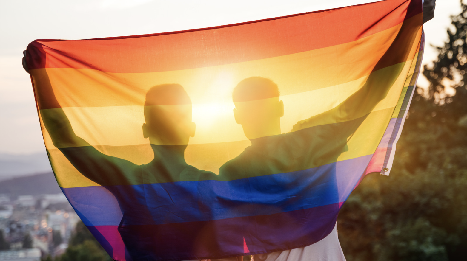 Two people holding a rainbow pride flag with sunlight shining through, symbolizing LGBTQ+ pride.