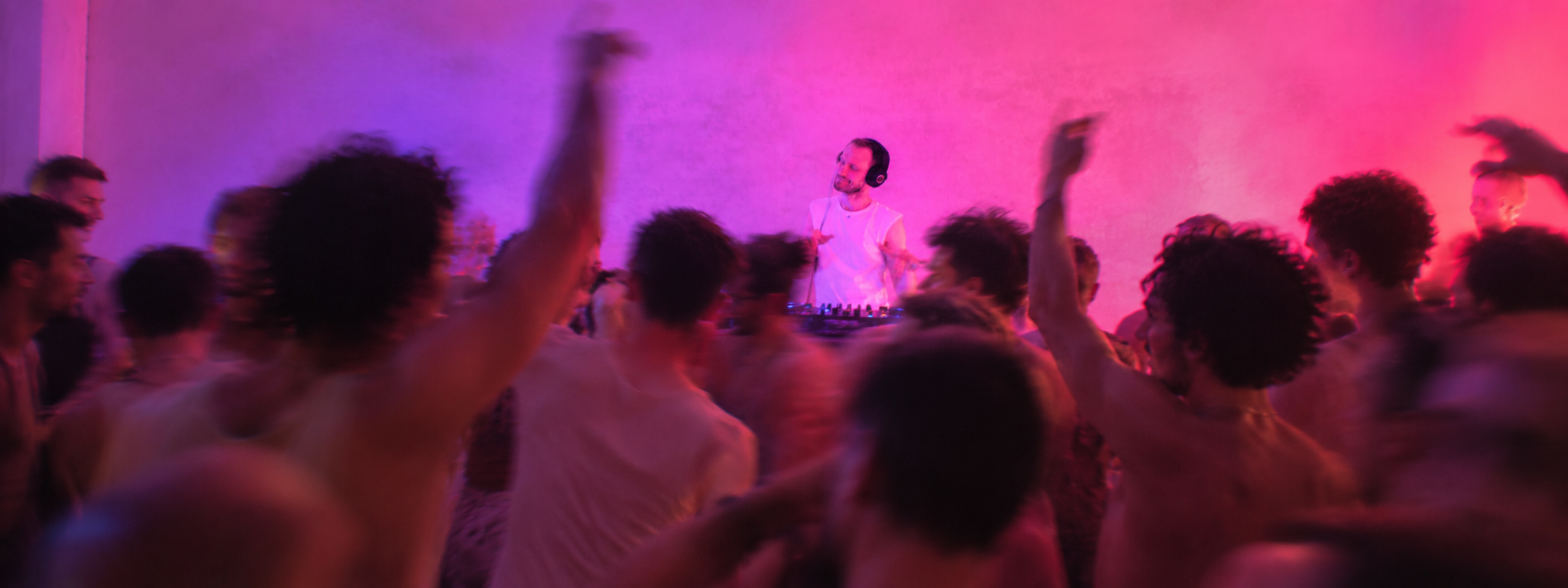 A DJ in a white sleeveless shirt wearing headphones, standing behind a DJ console, is surrounded by a crowd of people with their hands up, dancing in a dimly lit room with pink and purple lighting.