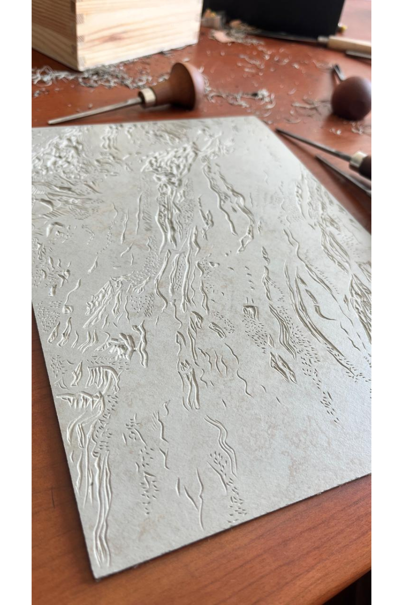 Close-up of a hand-carved linoleum block on a wooden studio desk with Pfeil mushroom-handled carving gouges and lino shavings in the background