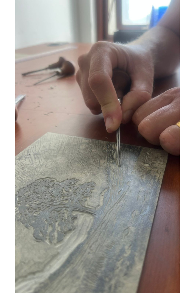 Close-up of an artist's hand using a carving tool to create intricate textures on a linoleum block, depicting a landscape with a tree in a home studio setting.