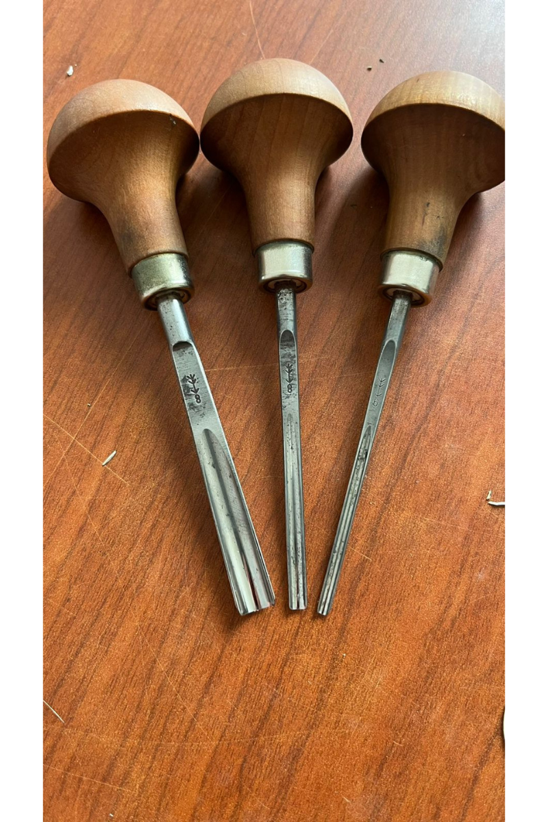 A top-down view of three professional Pfeil linocut carving gouges on a wooden desk, featuring ergonomic pearwood mushroom handles and various V-shaped and U-shaped steel tips.