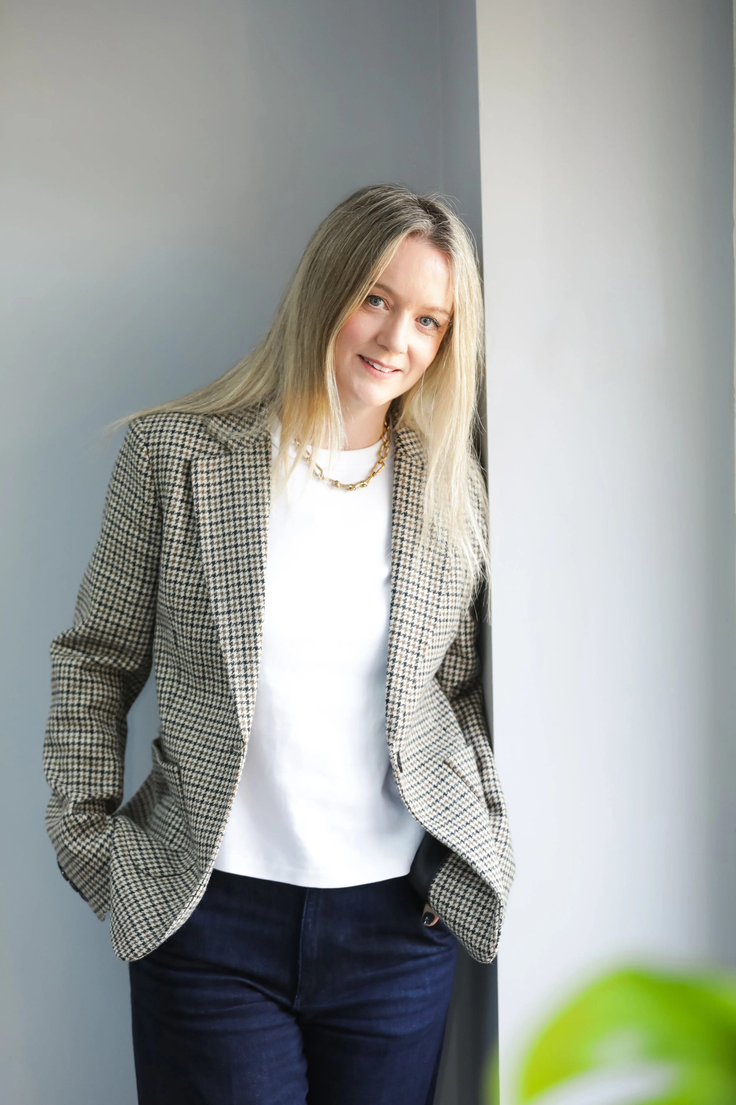 A woman with long blonde hair, wearing a checkered blazer, white top, and dark jeans, standing by a window with a smile.
