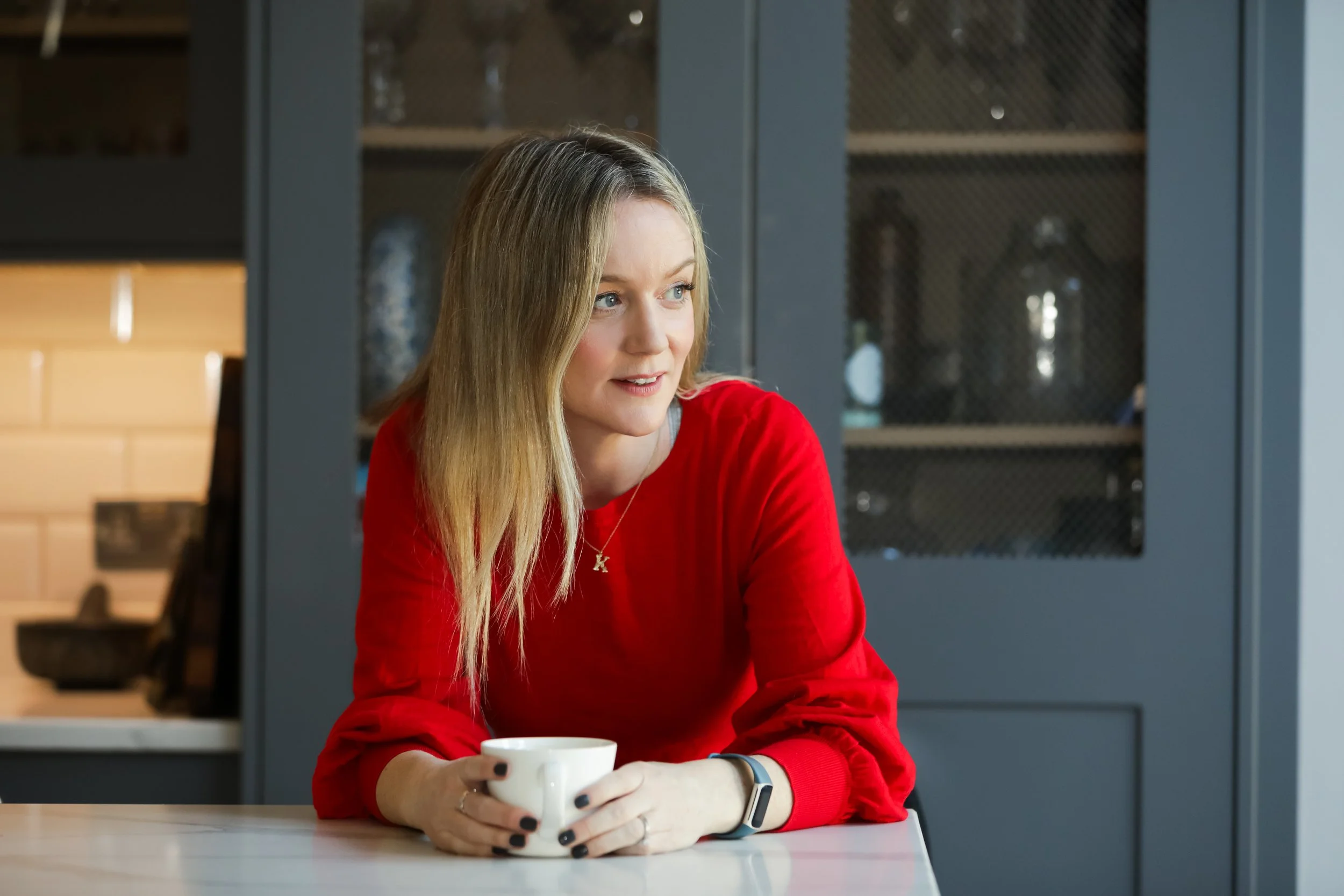 A woman with blonde hair wearing a red sweater, sitting at a white table, holding a white coffee cup, looking to her left in a modern kitchen with closed cabinets in the background.
