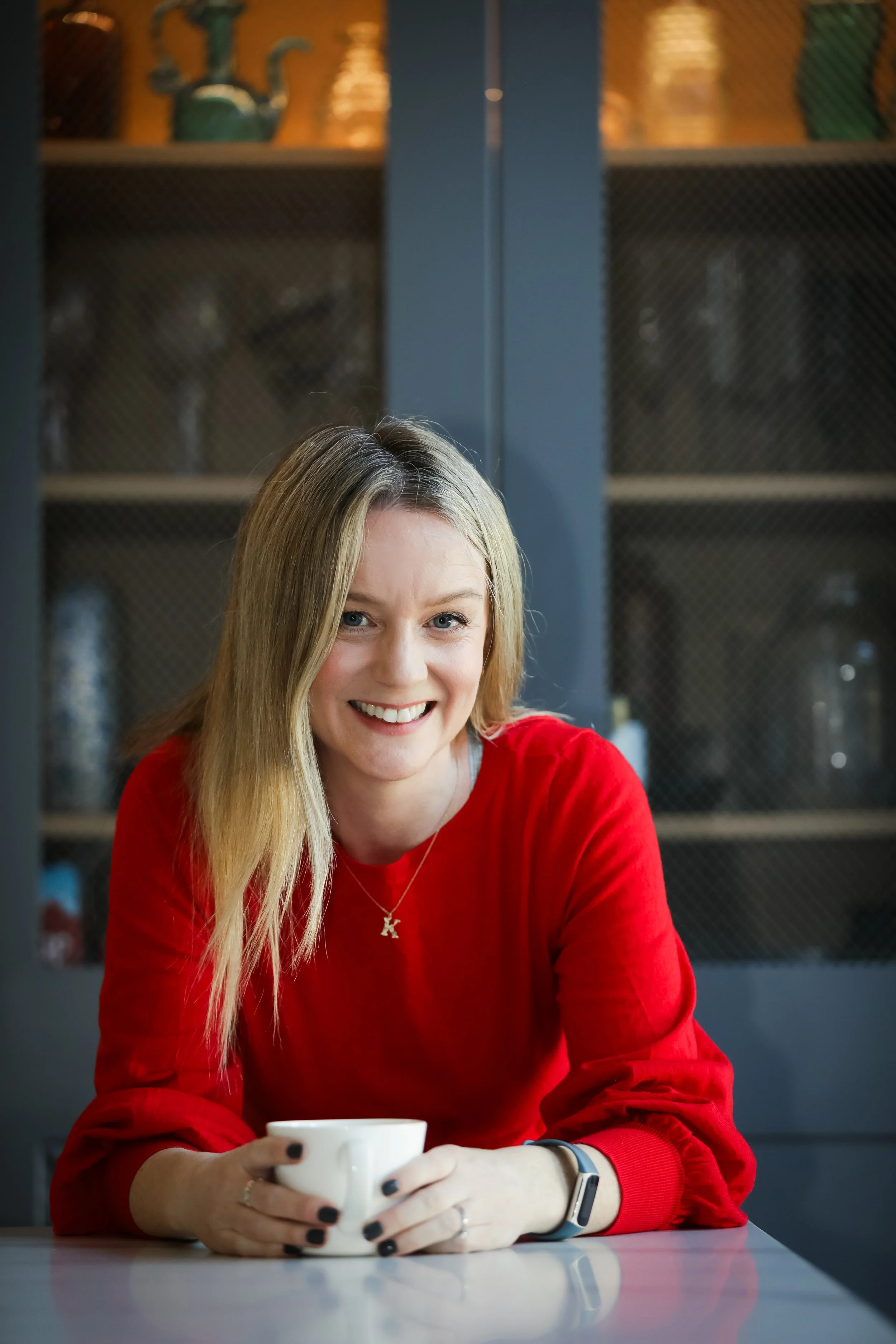 A woman with blonde hair wearing a red sweater sitting at a table, holding a white mug, smiling at the camera, with dark cabinets and glassware in the background.