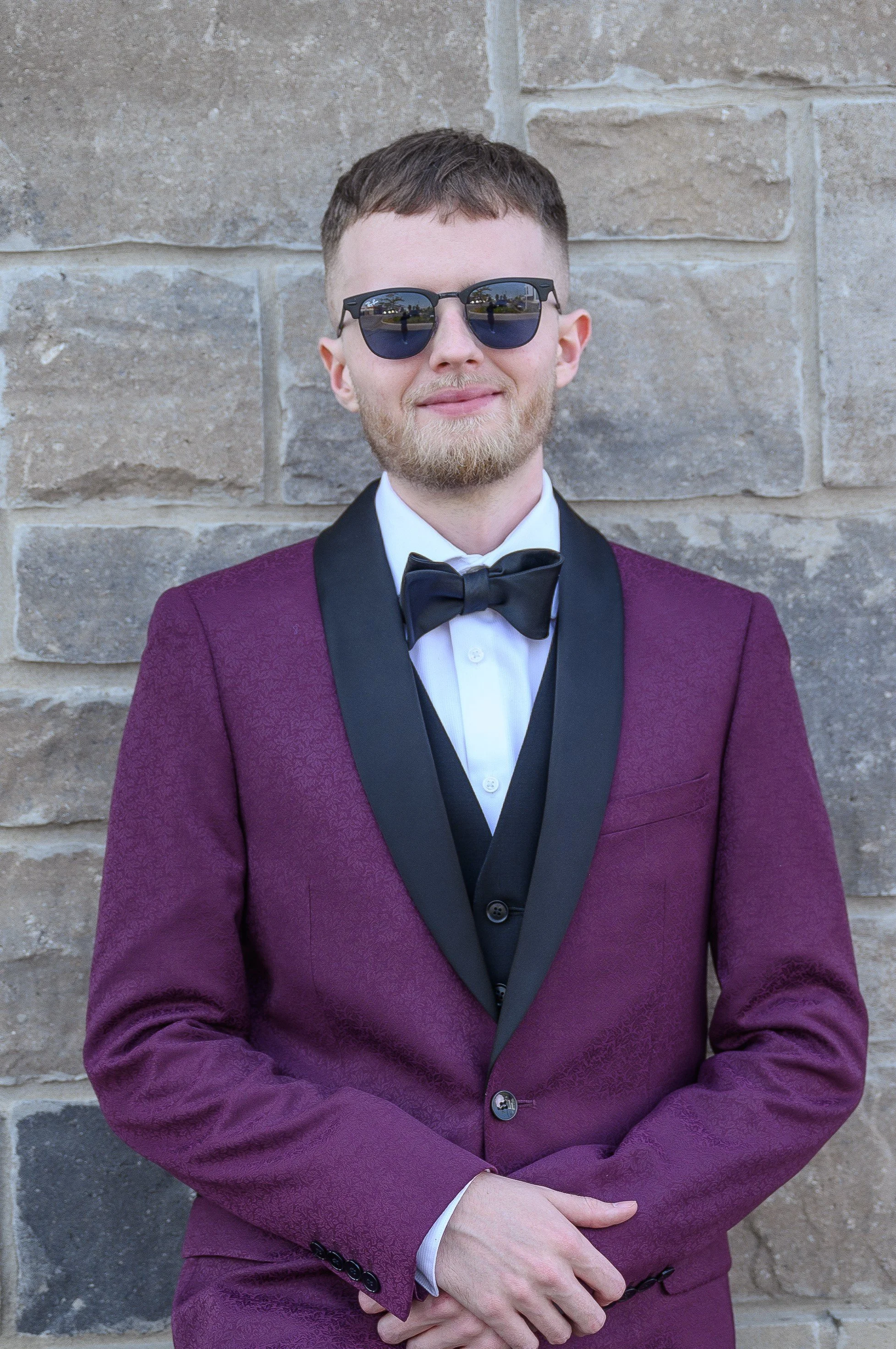 A young man in a purple tuxedo with a black lapel, white dress shirt, and black bow tie, standing in front of a stone wall, wearing sunglasses, and smiling.