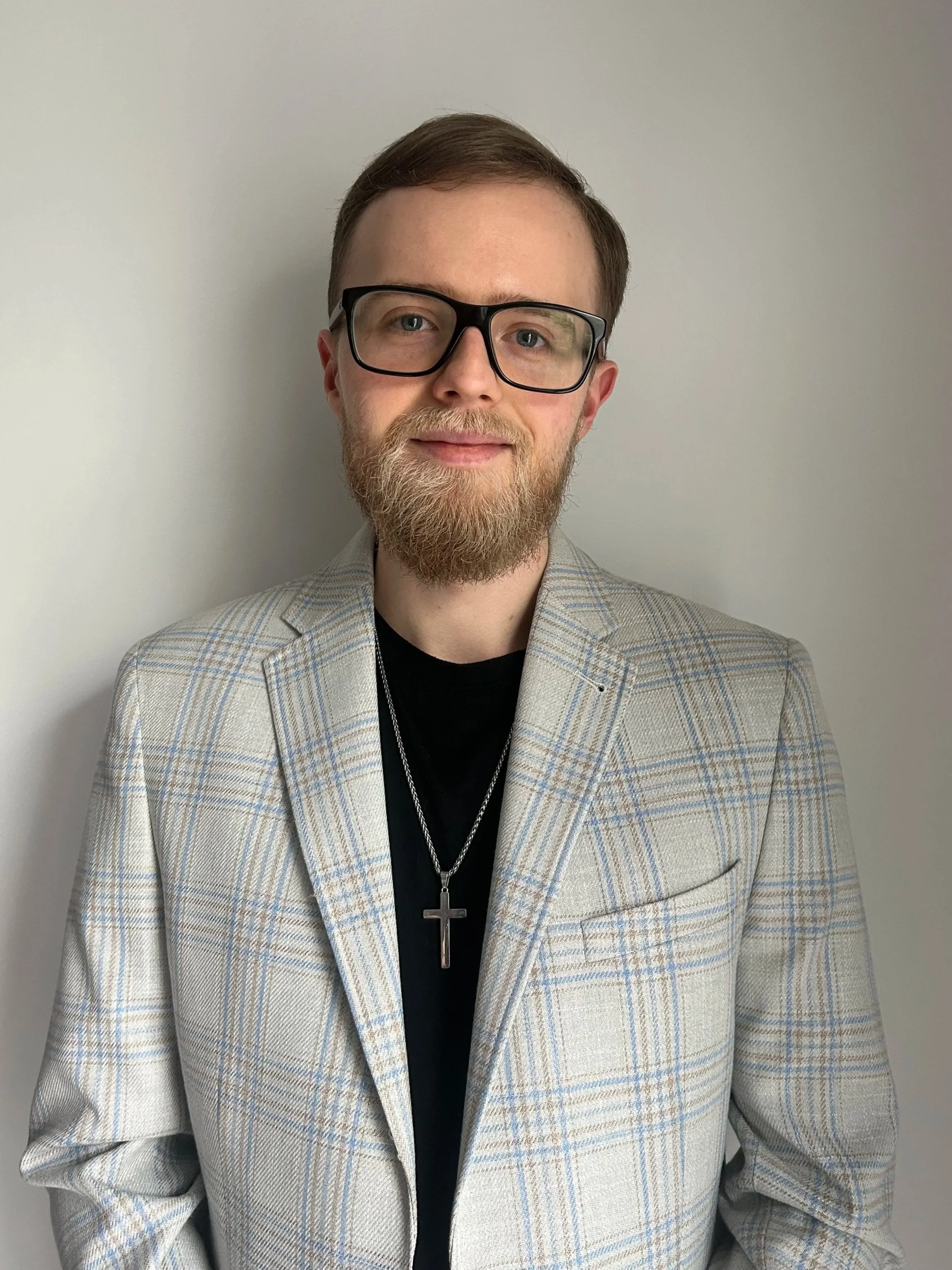 A man with glasses, a beard, and a cross necklace, wearing a plaid blazer over a black shirt, posing against a plain white wall.