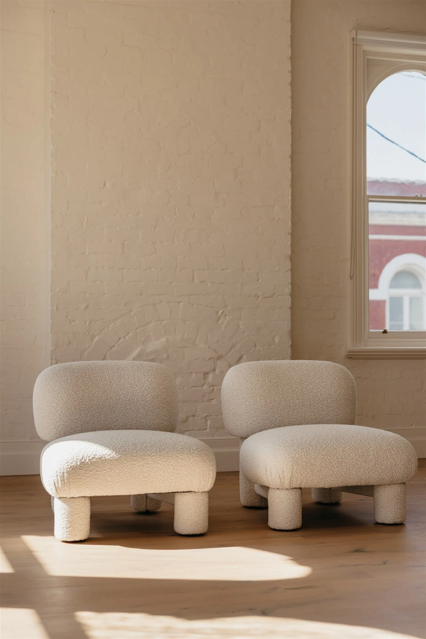 Two modern cream-colored upholstered chairs with rounded backs and cylindrical legs, placed inside a room with a wooden floor, a white brick wall, and a window showing an exterior building with arched windows.
