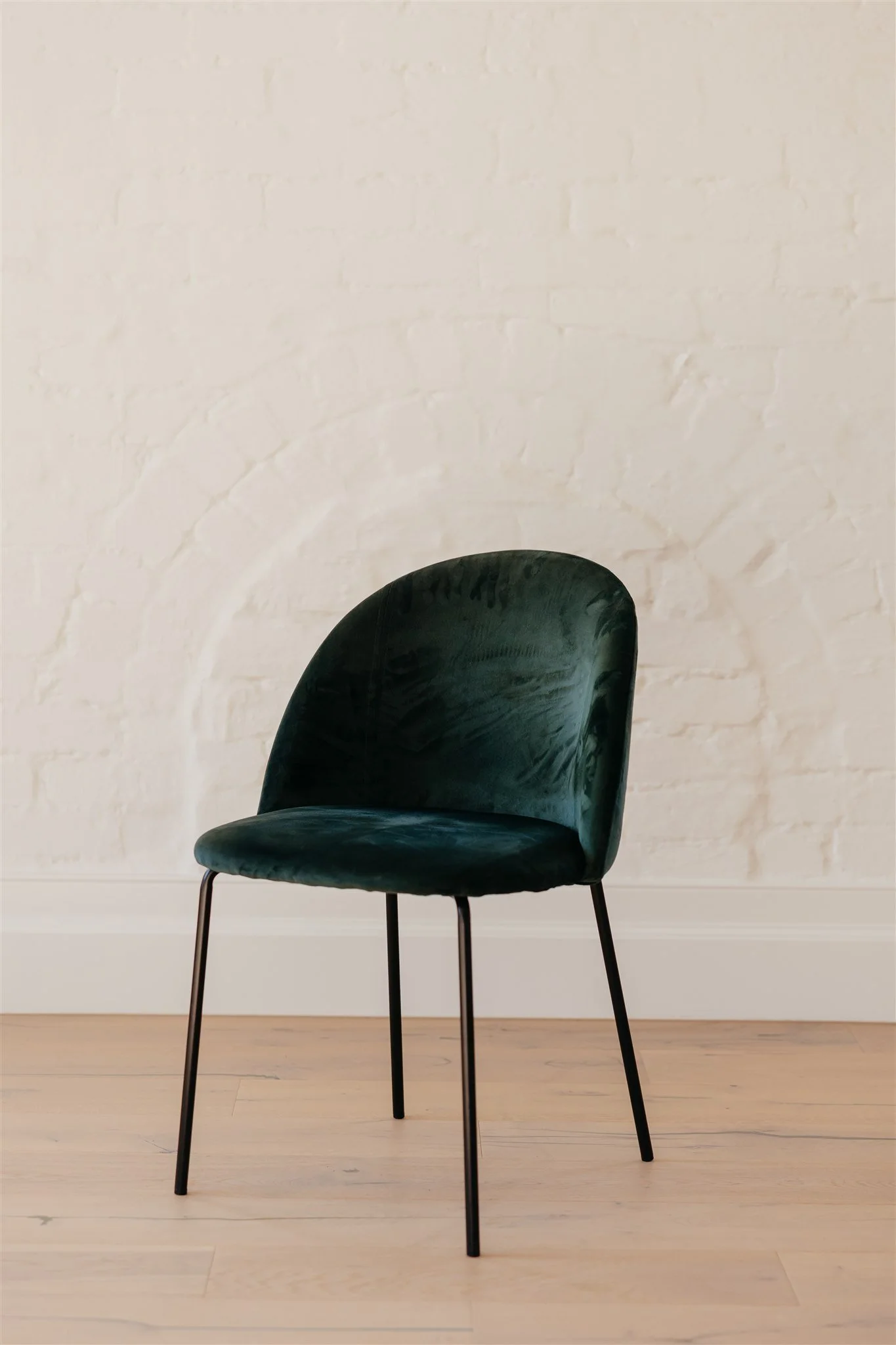 A dark green velvet chair with black metal legs against a white brick wall and light wood floor.