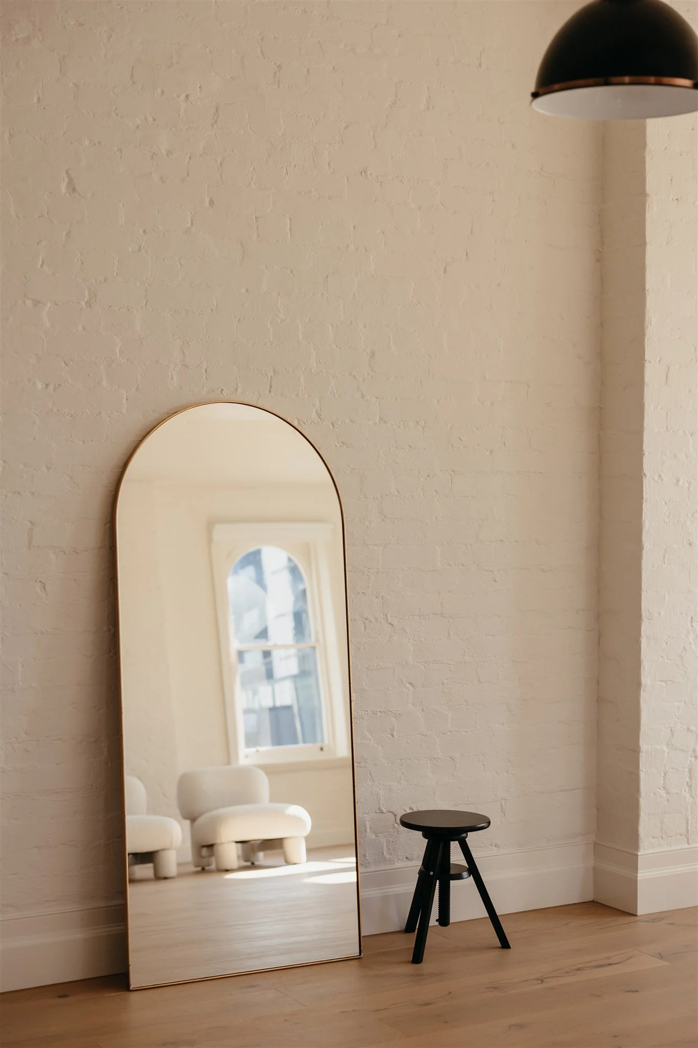 Full-length mirror leaning against a white brick wall, reflecting a window and two white chairs, with a black stool next to it on a wooden floor.