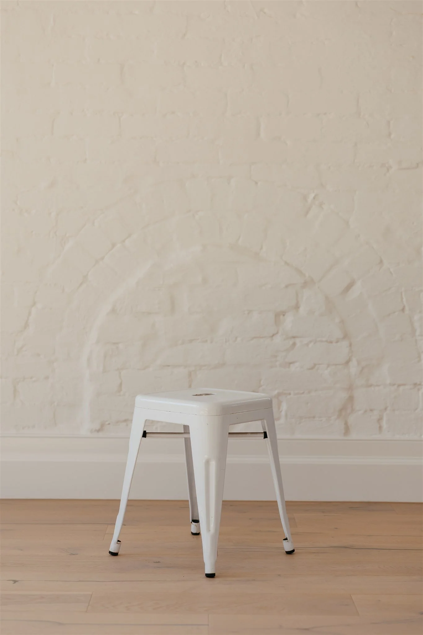 White metal stool with four legs on a wooden floor against a white brick wall