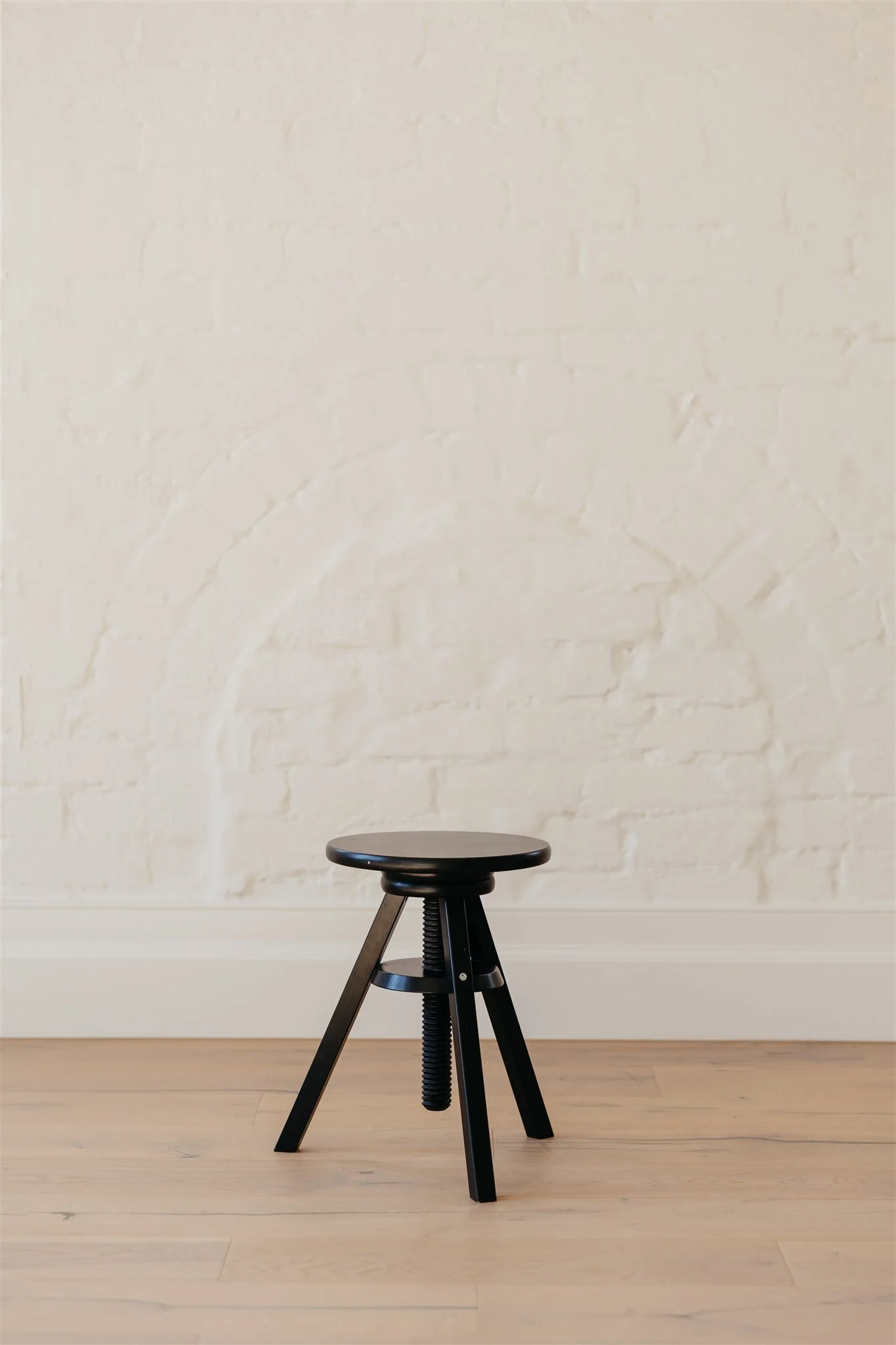 A small black adjustable stool with a round seat, set against a white brick wall and light wood floor.
