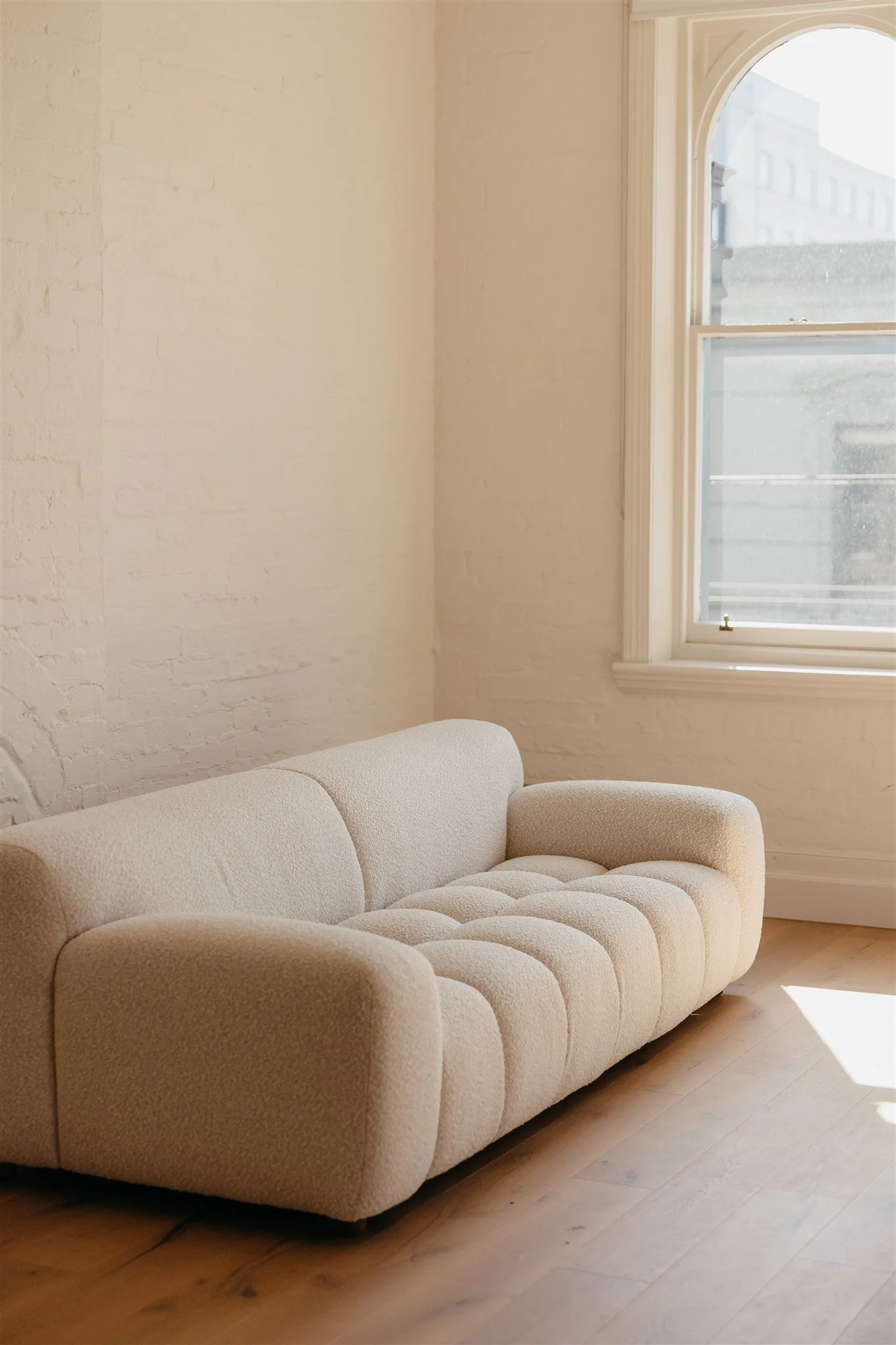 A cream-colored tufted sofa placed on a wooden floor next to a large window in a bright room with white brick walls.