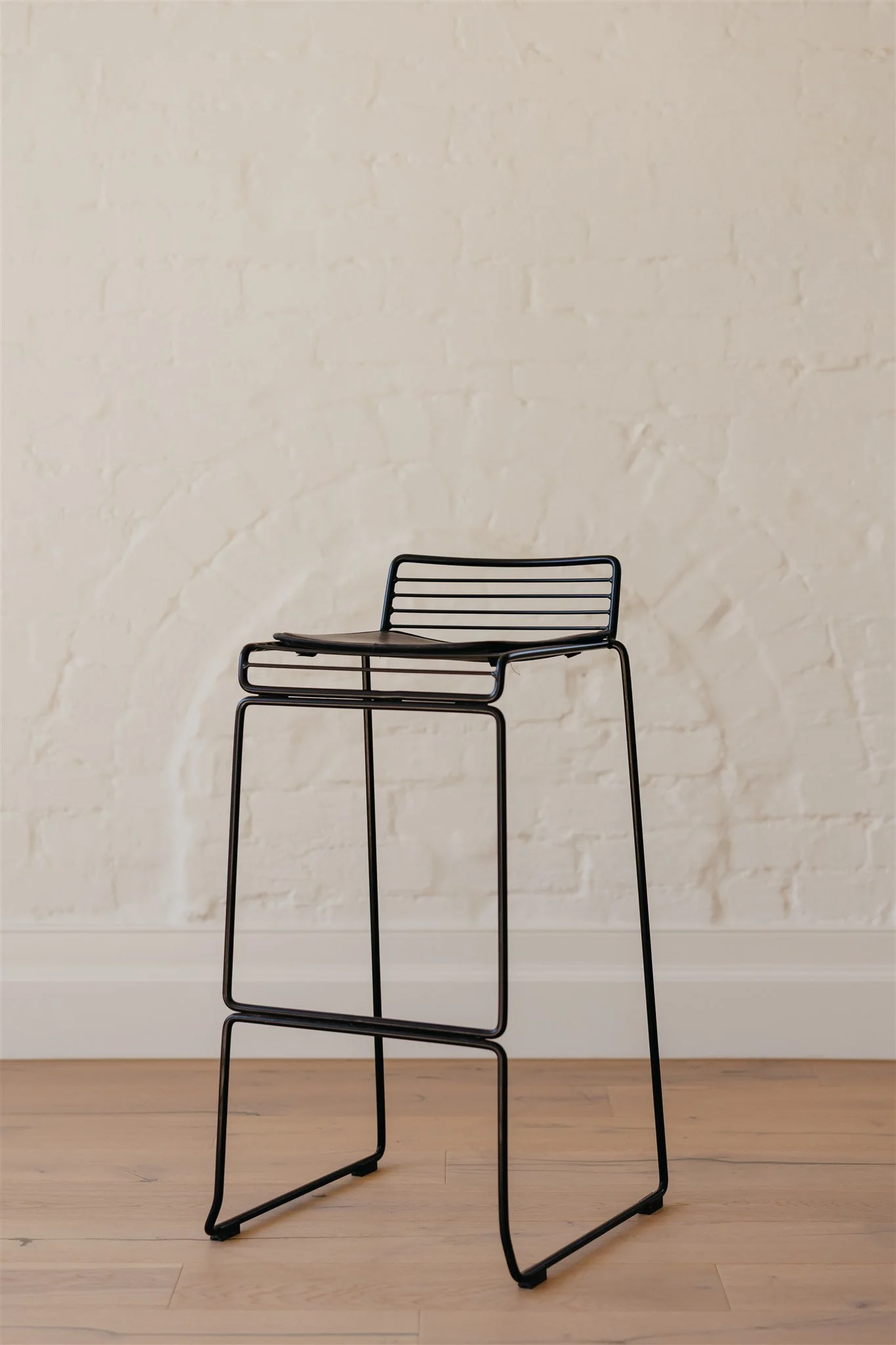 A black, modern wire-frame chair with a curved seat and a backrest placed against a light-colored brick wall and wooden floor.