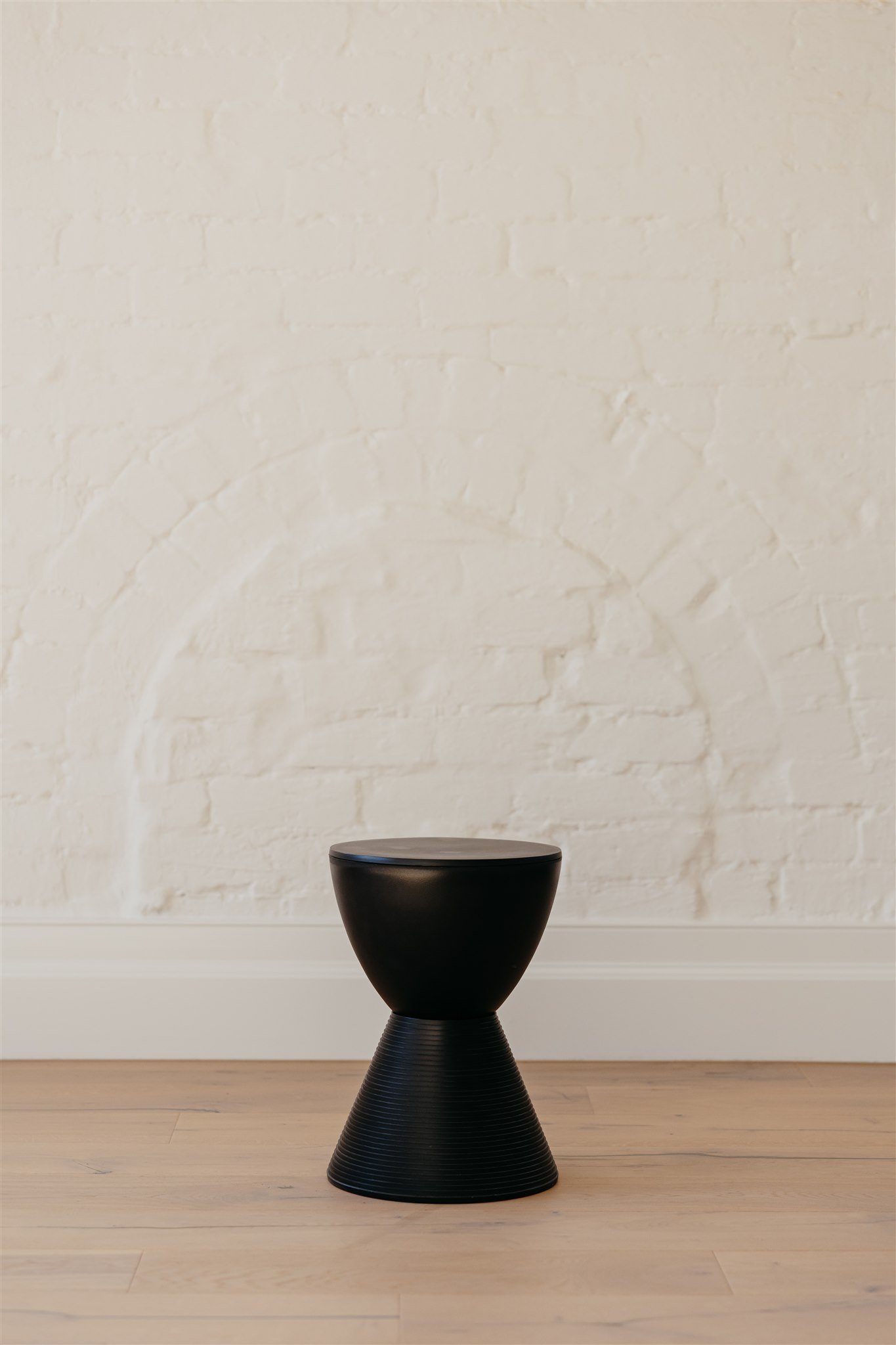 A minimalist black stool with a conical base and rounded seat against a white brick wall and light wooden floor.