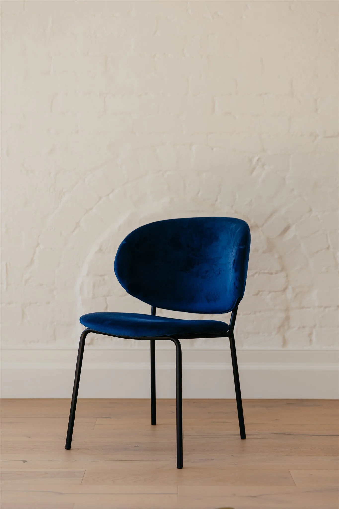 A modern chair with a dark blue velvet cushion and black metal legs, placed against a white brick wall and light hardwood floor.