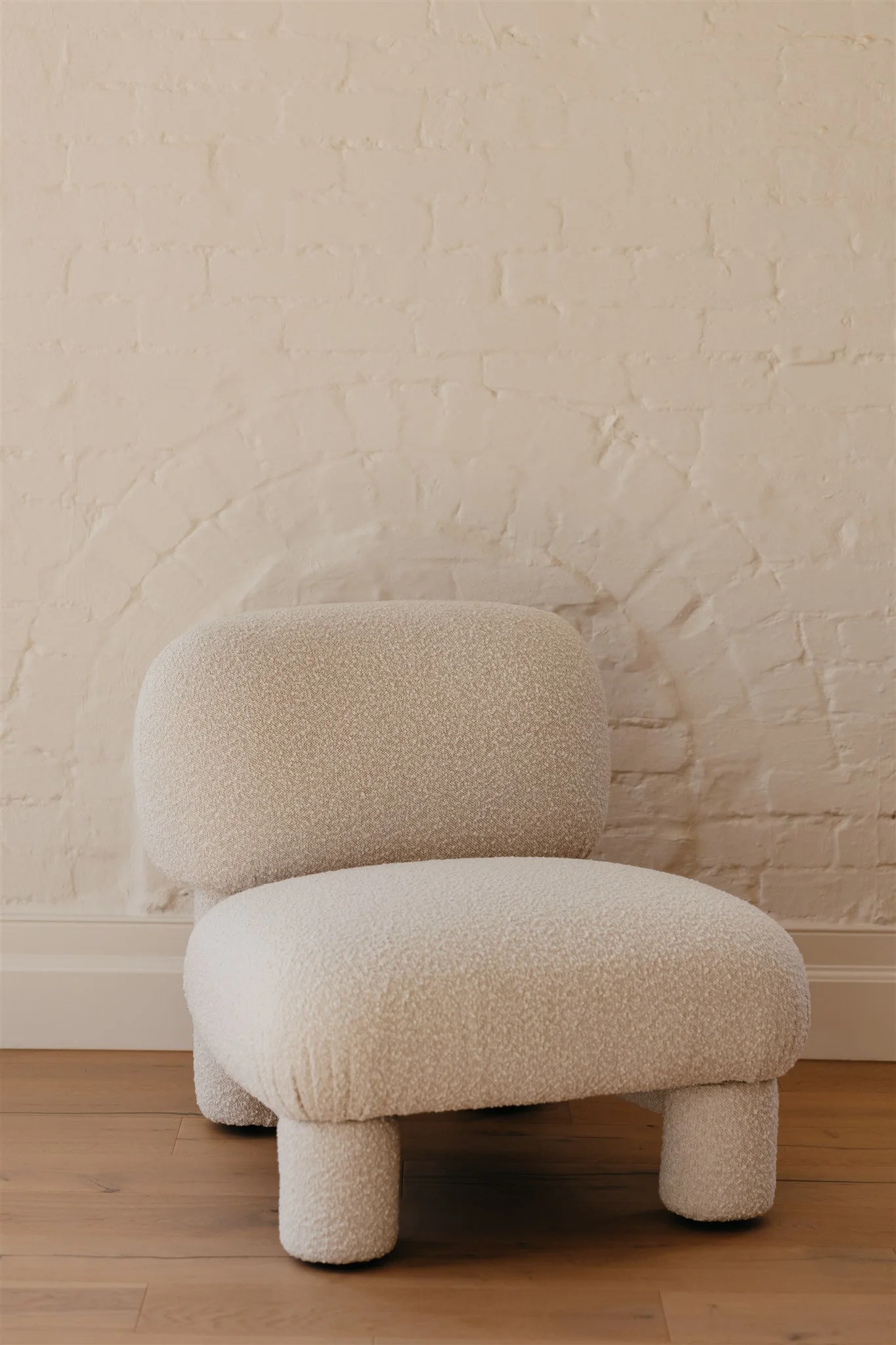 A modern, plush beige lounge chair with a rounded back and four short legs in front of a white brick wall with an arch detail.