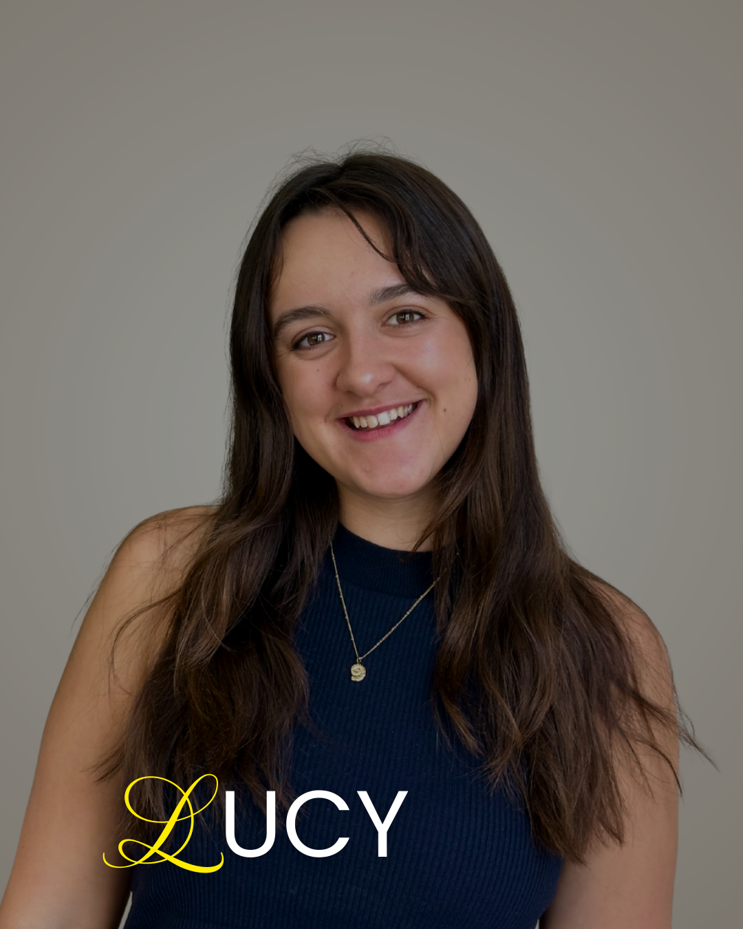 A young woman with long dark hair smiling at the camera, wearing a navy sleeveless top and a gold necklace with a small pendant, against a plain gray background. The word 'LUCY' is written in yellow and white text at the bottom left corner.