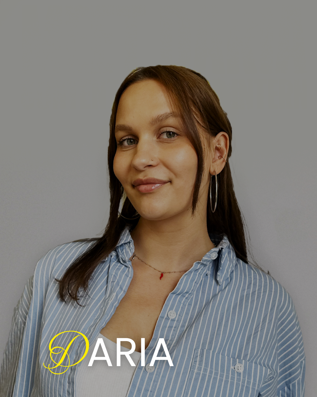 A woman with brown hair, blue eyes, and light skin, smiling slightly in front of a plain gray background. She is wearing a striped blue and white button-up shirt, hoop earrings, a small nose piercing, and a delicate necklace with a small red pendant. The name "Daria" is written at the bottom of the image.