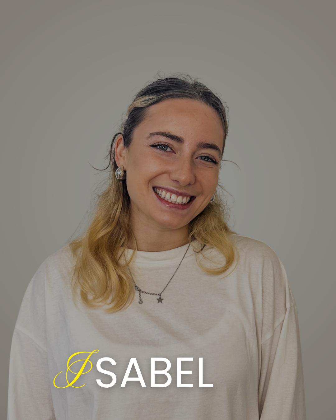 A young woman with blonde hair, blue eyes, and a cheerful smile, wearing silver earrings, a star-shaped necklace, and a cream-colored top, standing against a plain gray background.
