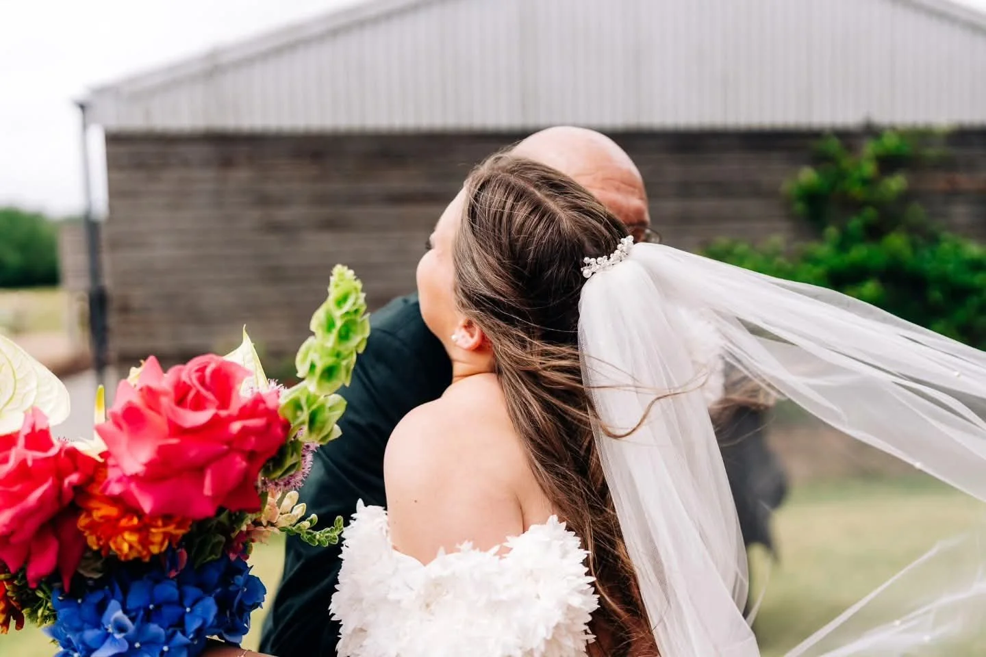 A bride and groom sharing a kiss outdoors, the bride wearing a white off-shoulder gown and veil, holding a colorful bouquet of flowers, with a rustic barn in the background.
