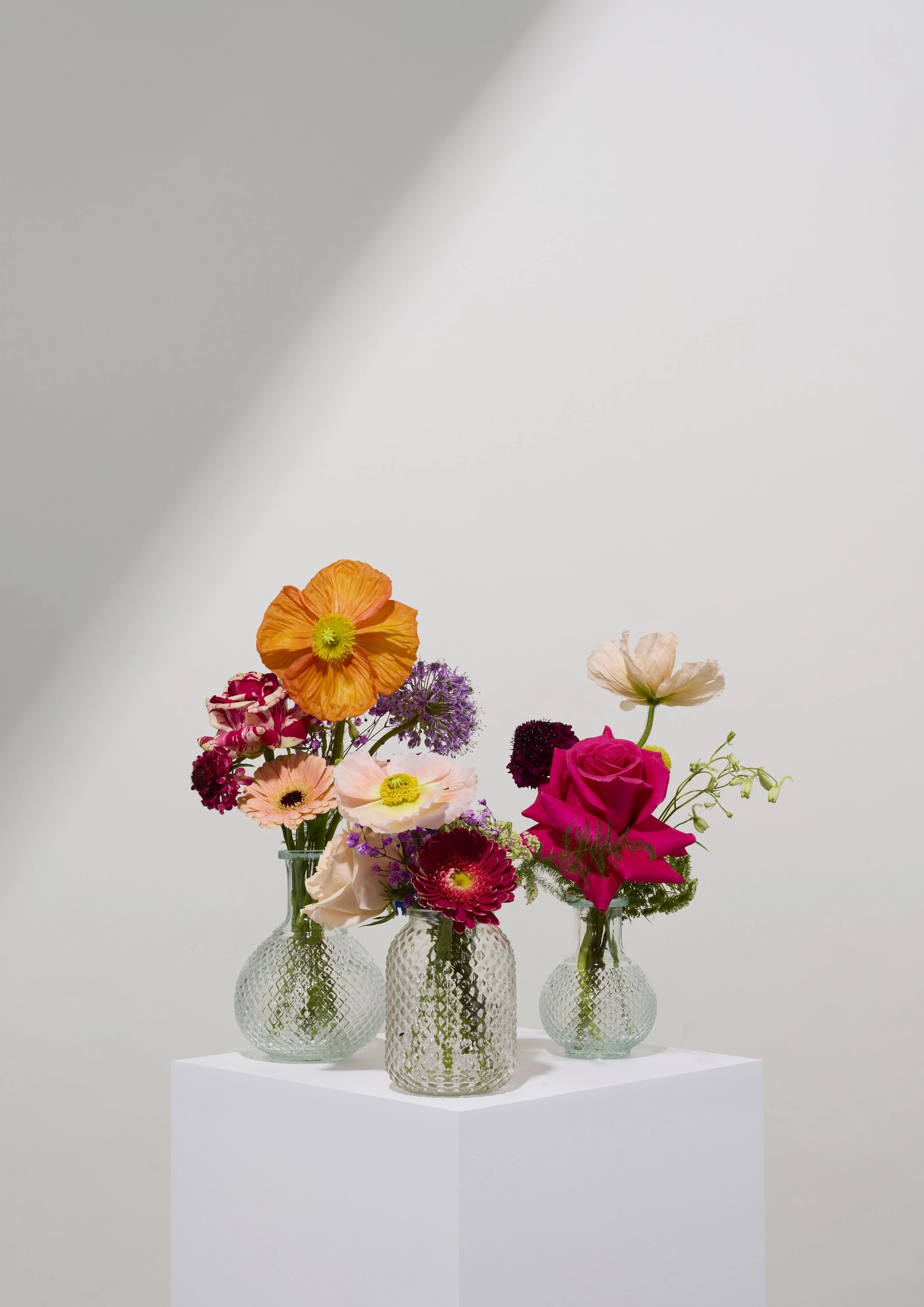 Three glass vases with colorful flowers on a white pedestal against a plain background.