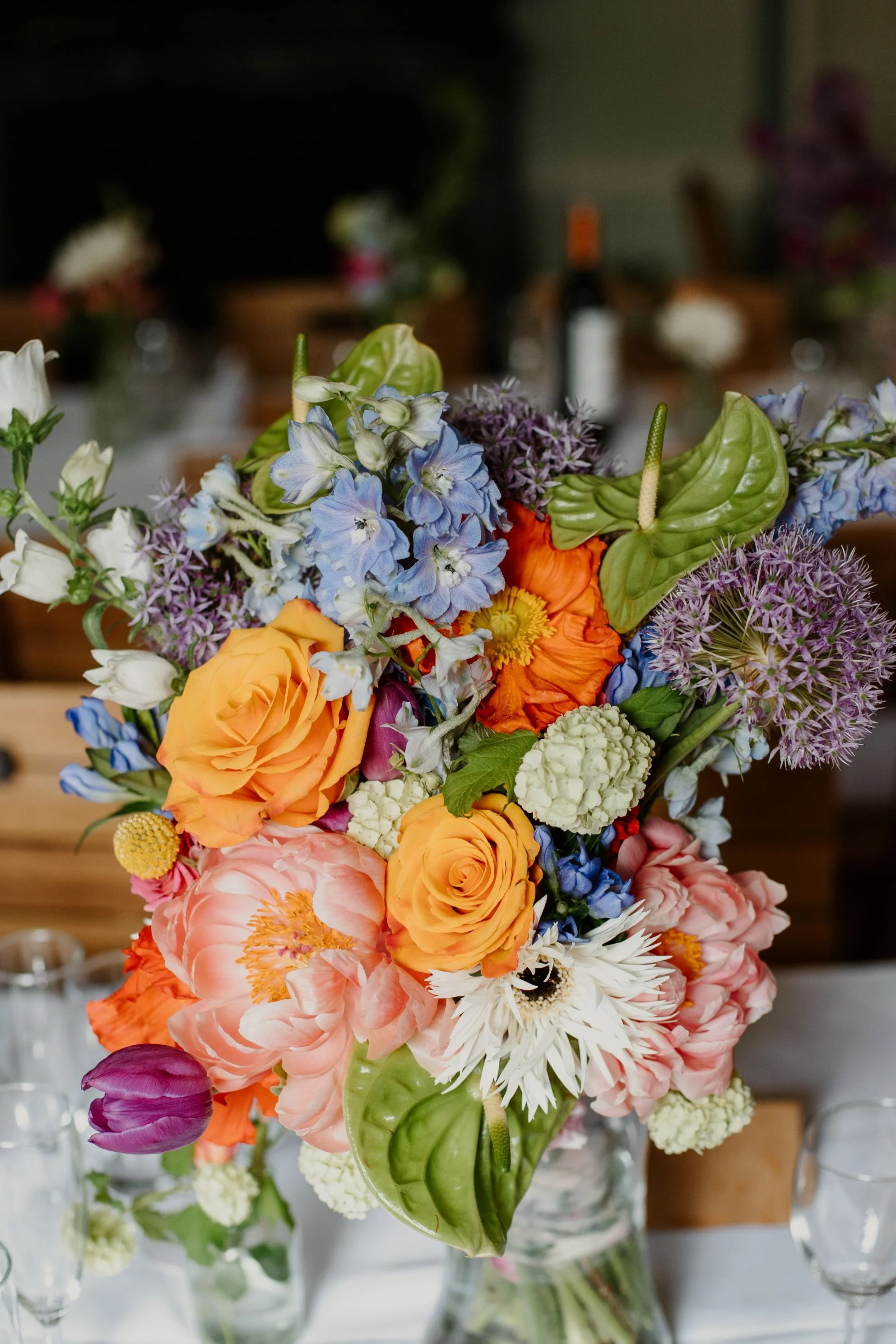 Colorful bouquet of various flowers including roses, tulips, anemones, and delphiniums in a glass vase on a table.