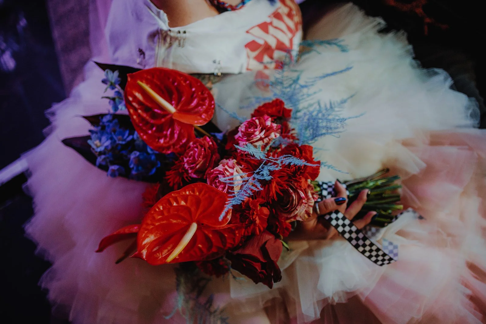Person in a colorful dress holding a bouquet of vibrant flowers including red anthuriums, pink roses, blue flowers, and decorative fern leaves.