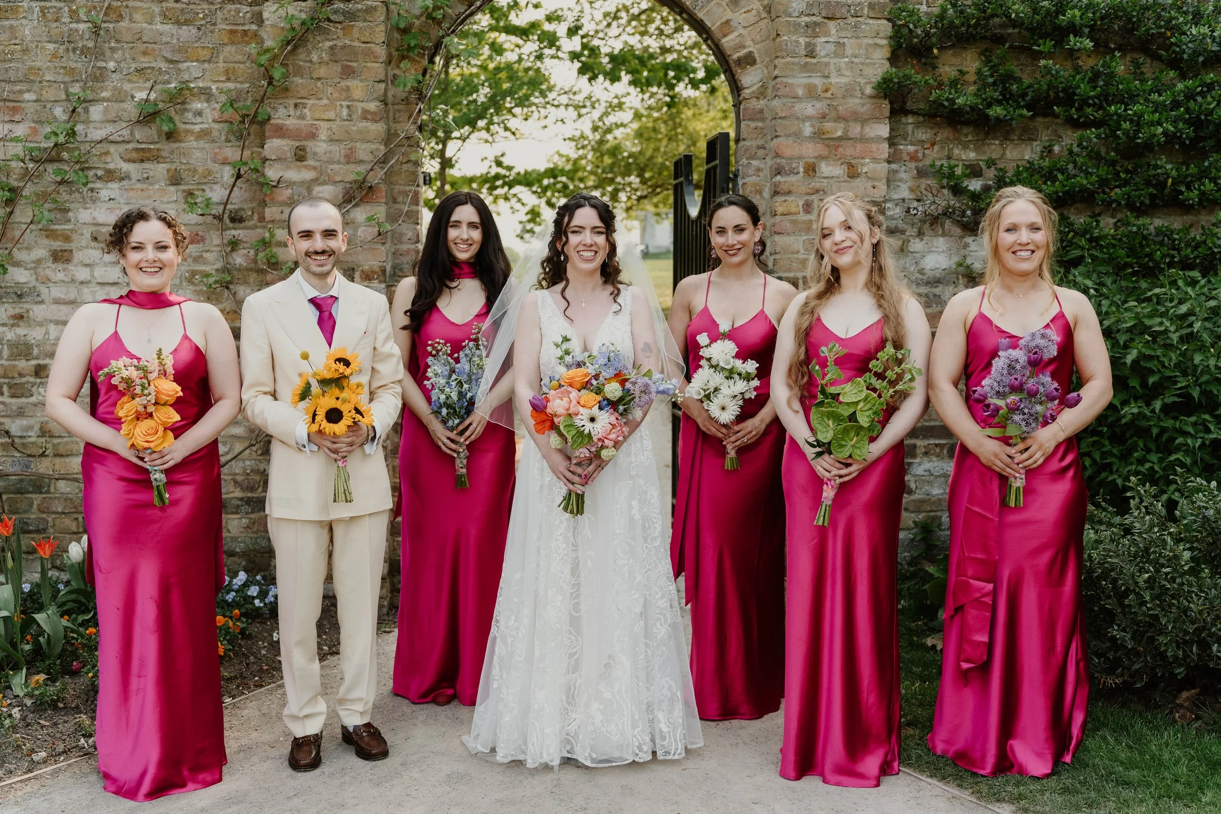 A wedding party standing outdoors in front of a brick archway, including a bride in a white lace dress with a veil, surrounded by six bridesmaids in matching hot pink satin dresses, all holding colorful floral bouquets, and a groom in a cream suit wi