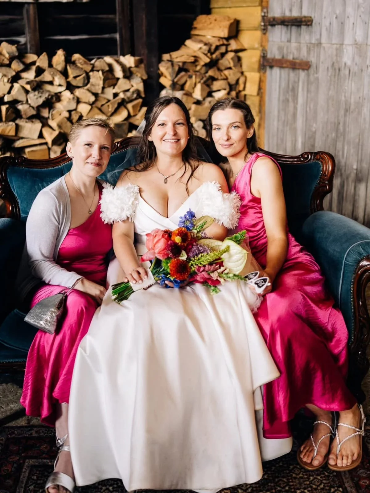 Three women sitting on a vintage blue couch, with a focus on the bride in the center holding a colorful bouquet, flanked by two women in pink dresses in a rustic setting with stacked firewood in the background.