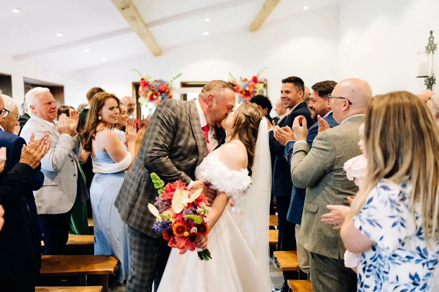Wedding ceremony with bride and groom kissing, surrounded by family and friends clapping and smiling indoors with floral decorations.
