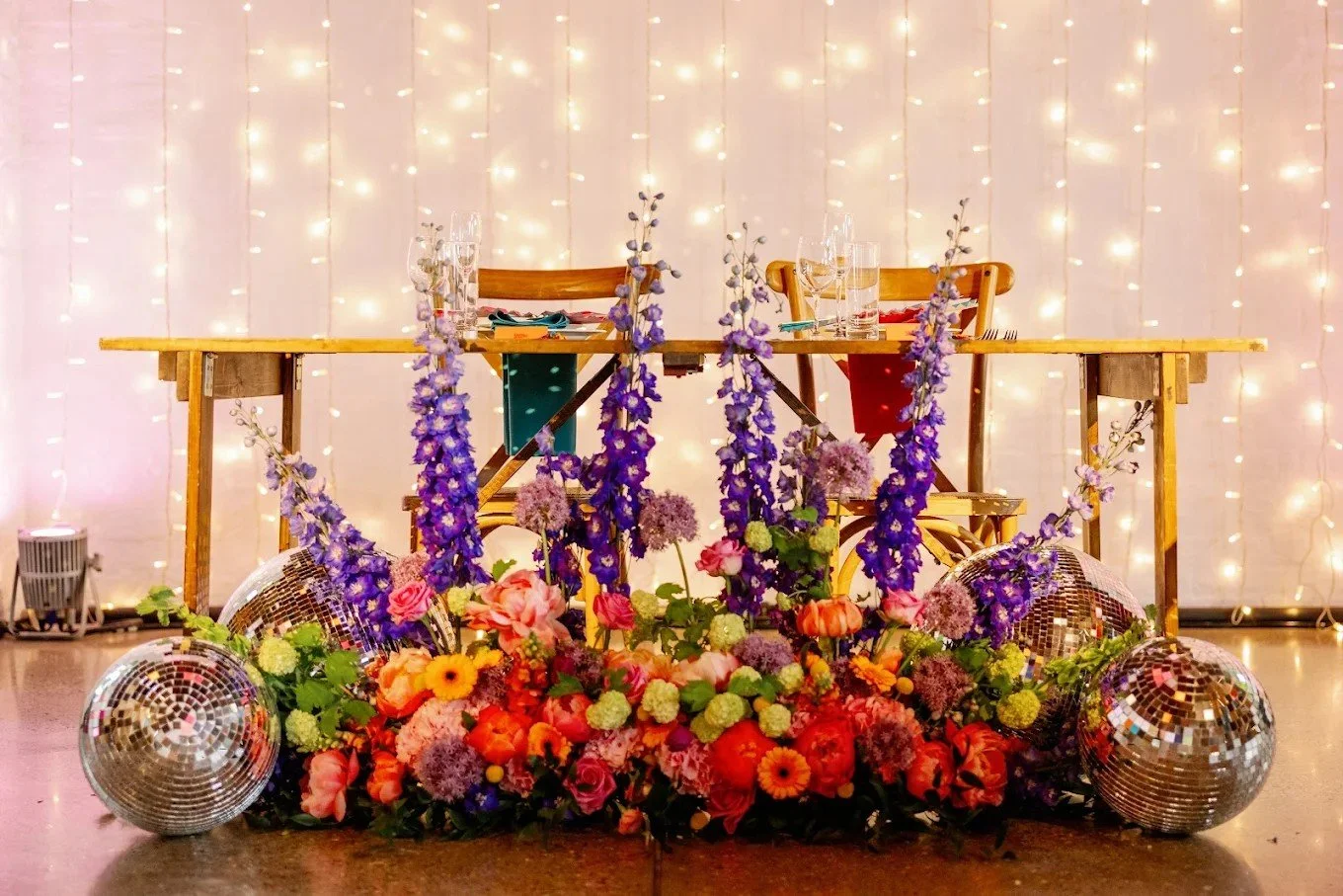 A decorated table setup with purple, pink, and yellow flowers, disco balls, and fairy lights in the background.