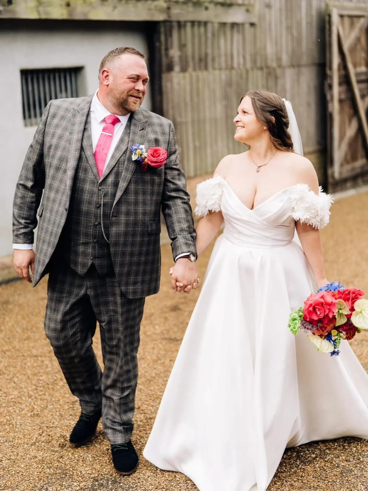A couple walking hand in hand outdoors, dressed in wedding attire, smiling at each other. The man is wearing a gray plaid suit with a pink tie and boutonniere, and the woman is in a white wedding dress with off-the-shoulder fluffy sleeves, holding a 