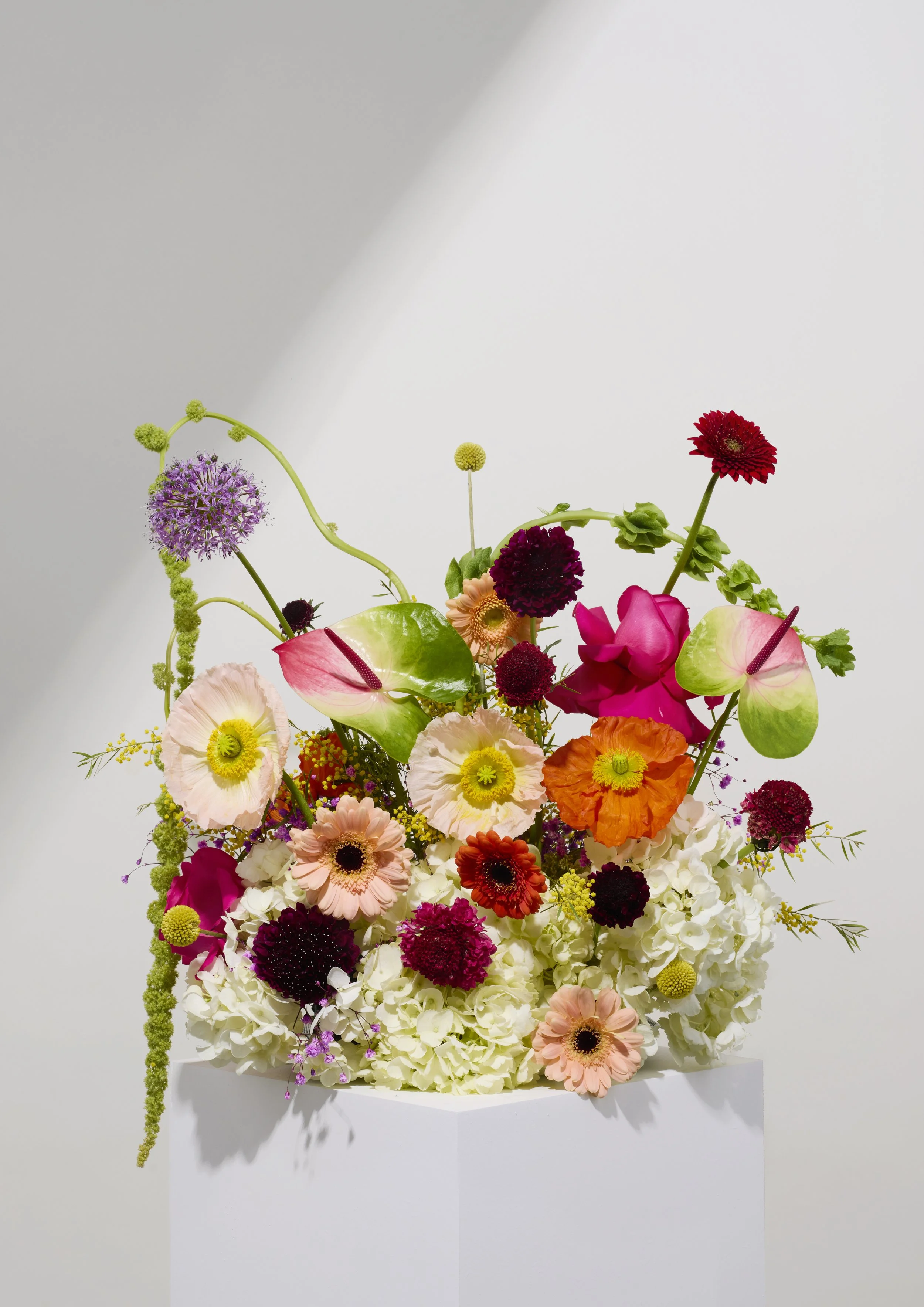 Colorful flower arrangement with pink, red, purple, orange, and white flowers in a white vase on a plain white background.
