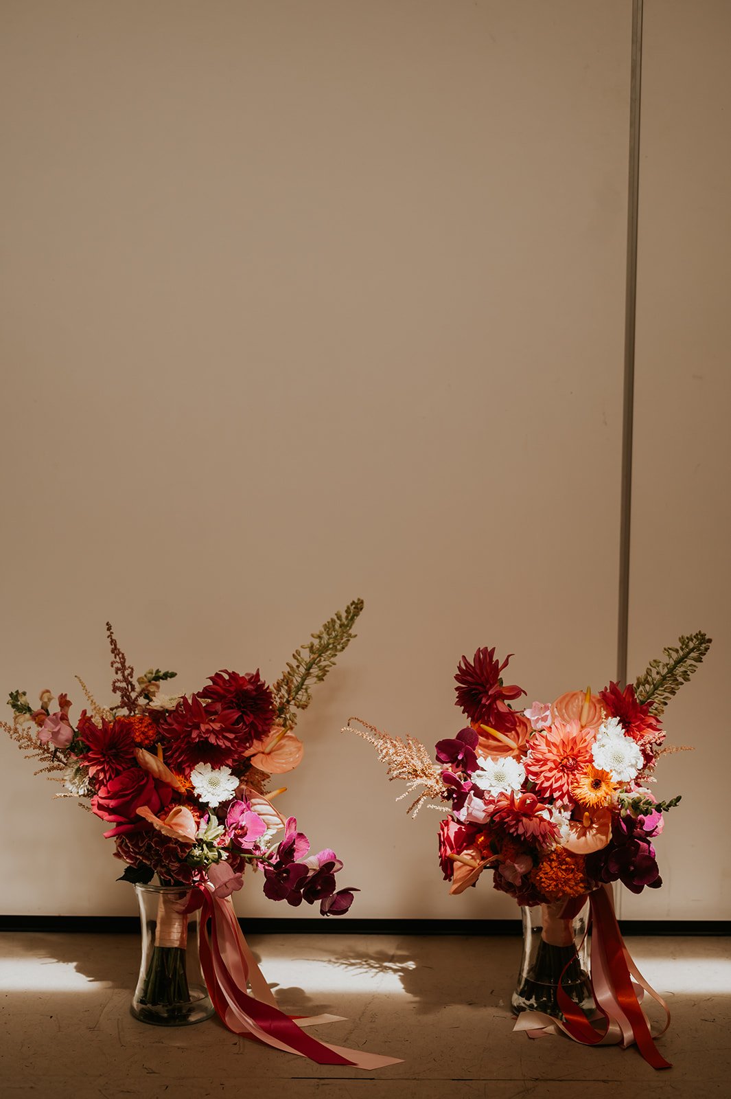 Two floral bouquets in glass vases with pink and red flowers, ribbons, and greenery, placed on the floor against a plain wall.
