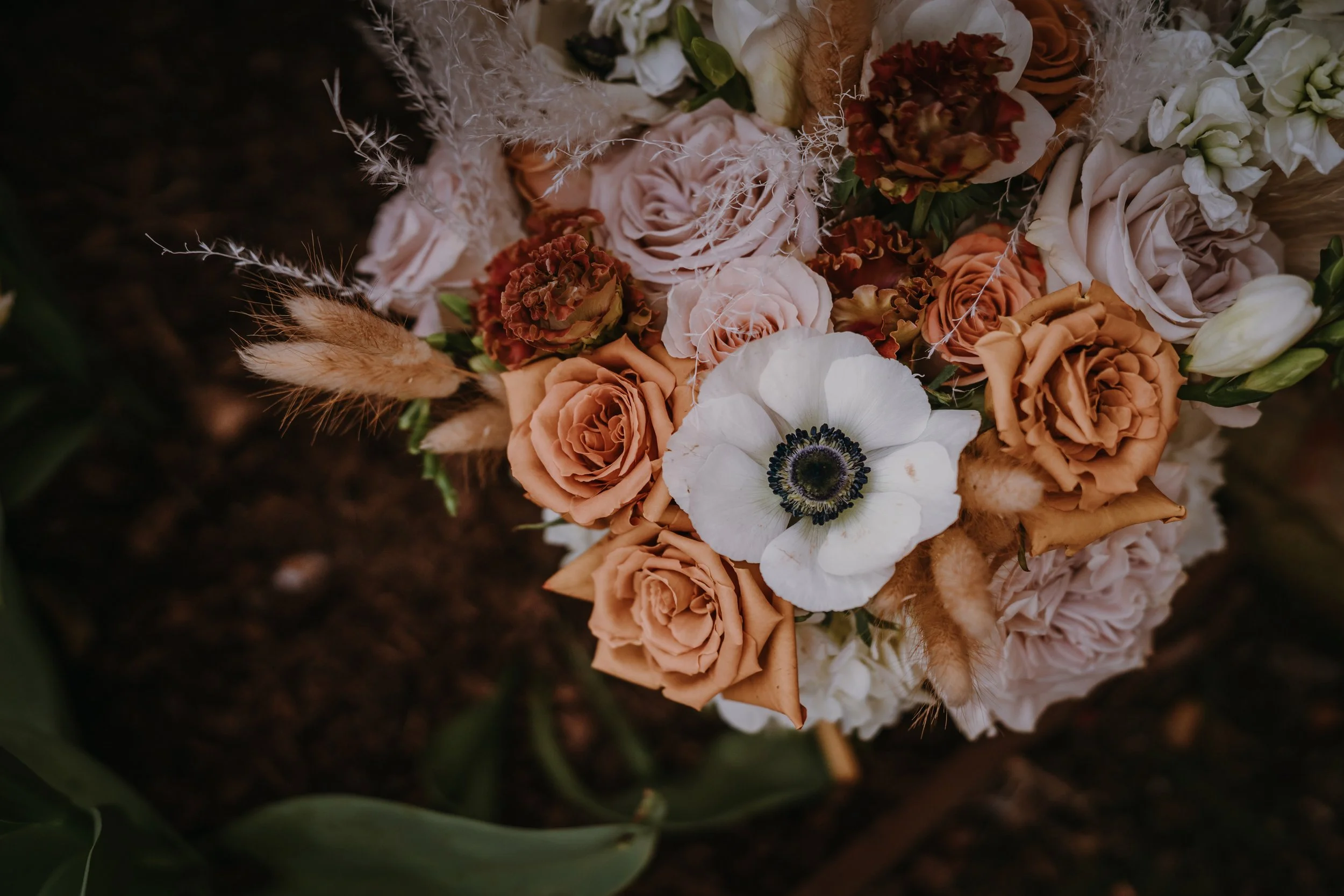 Close-up of a flower arrangement with roses, anemones, and dried grasses on dark soil background.