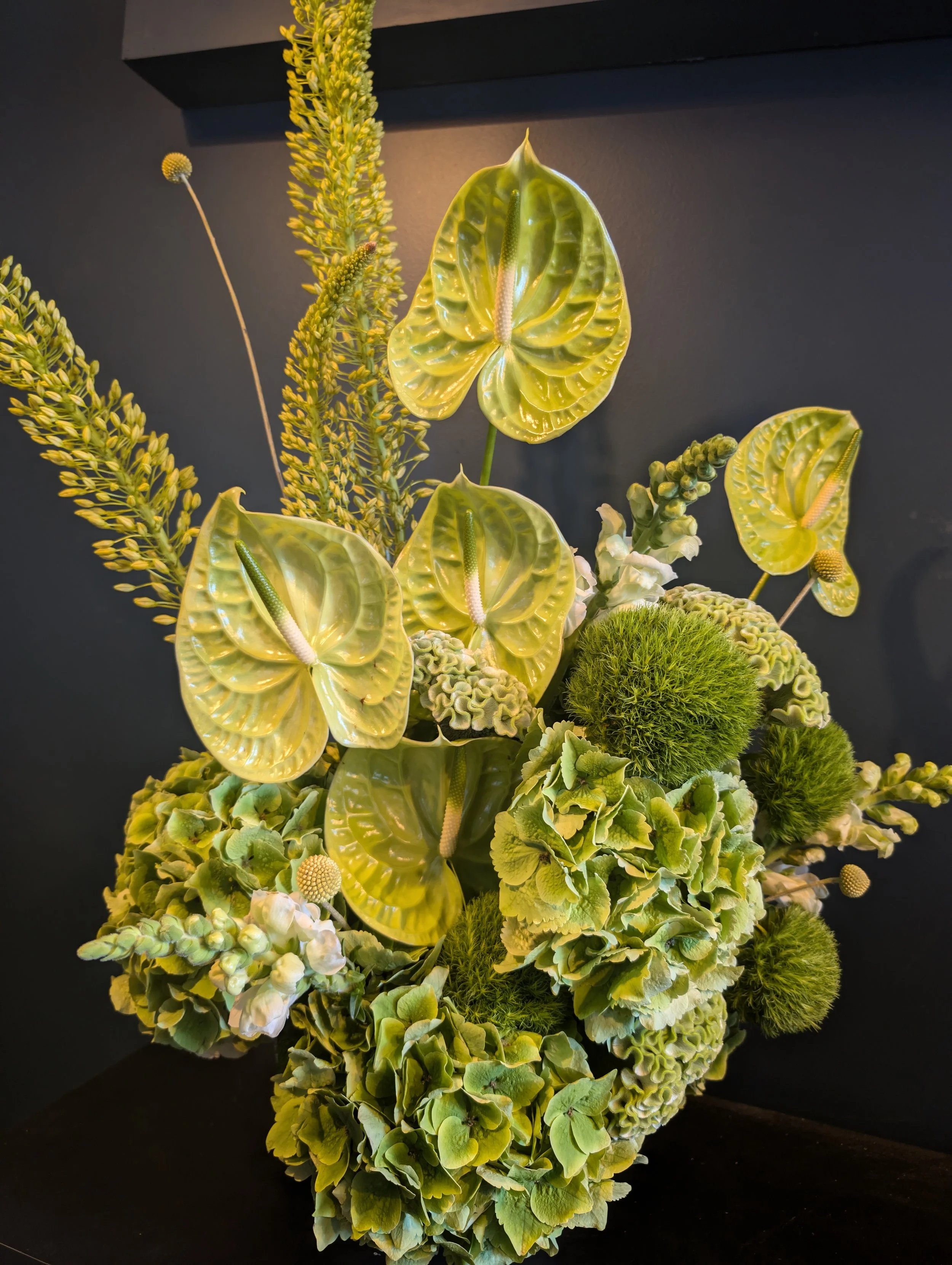 Artificial floral arrangement with green anthuriums, bright green ball-shaped moss, green hydrangea, and additional green foliage set against a dark background.