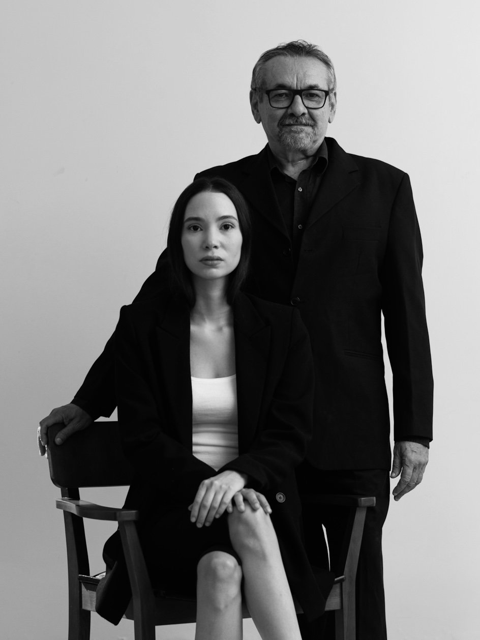 Homem mais velho de cabelo e barba grisalhos, usando óculos, vestindo terno preto, e mulher mais jovem de cabelo escuro, usando blazer preto e camiseta branca, sentado em cadeira com a mão sobre o braço e mulher de pé ao lado, em um fundo branco.