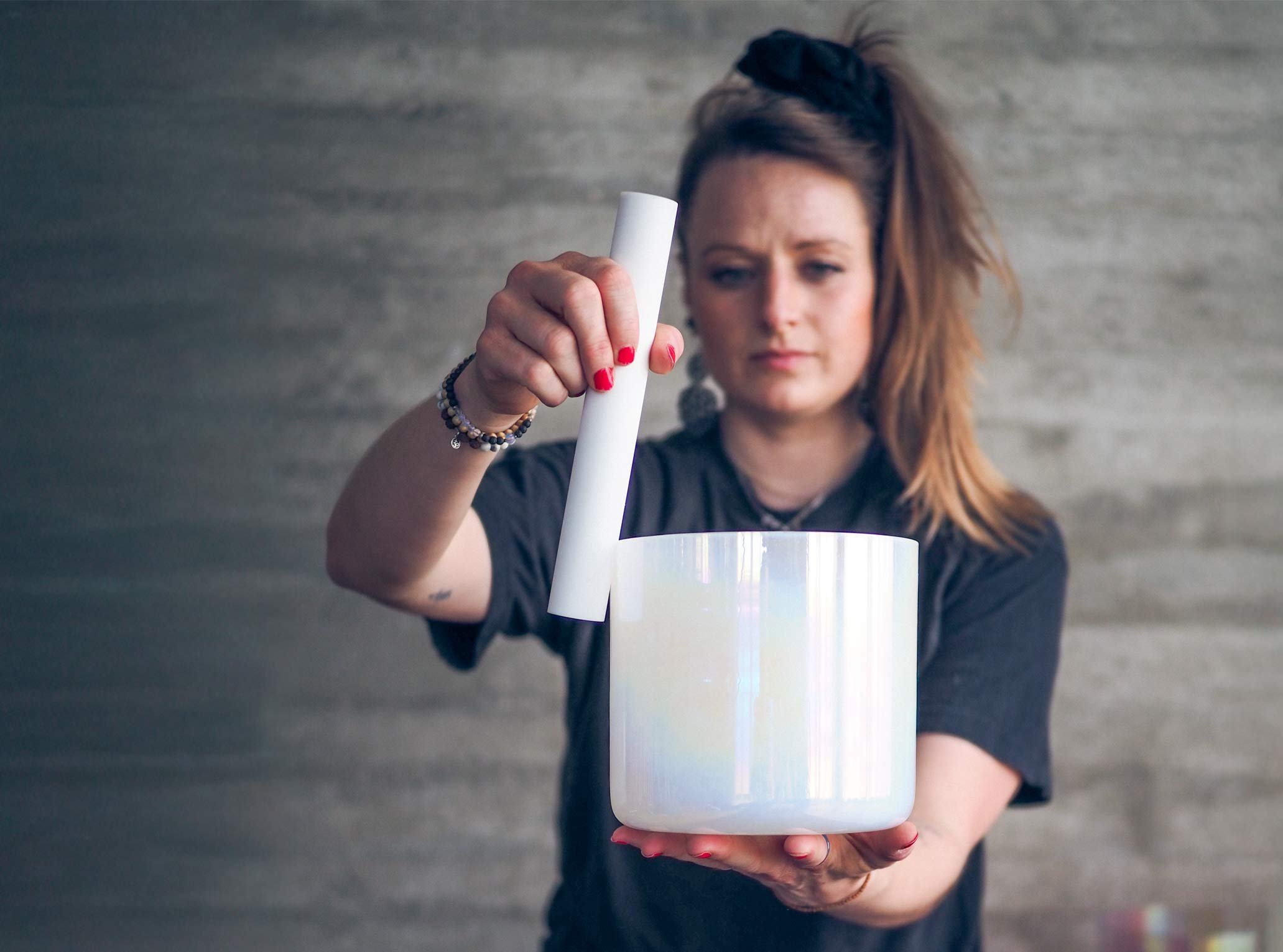 Eine Frau hält eine milchig weiße Kristallklangschale in die Höhe und bespielt diese auf ihrer Handfläche mit einem Schlegl