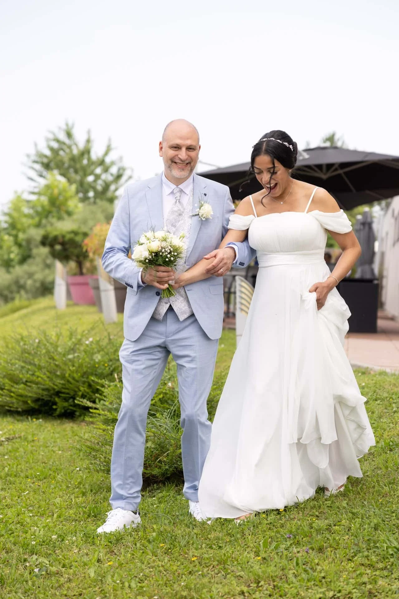 Un couple de mariés marche dans un jardin lors de leur mariage, la femme en robe blanche et l'homme en costume bleu clair, tenant une bouquet de fleurs.