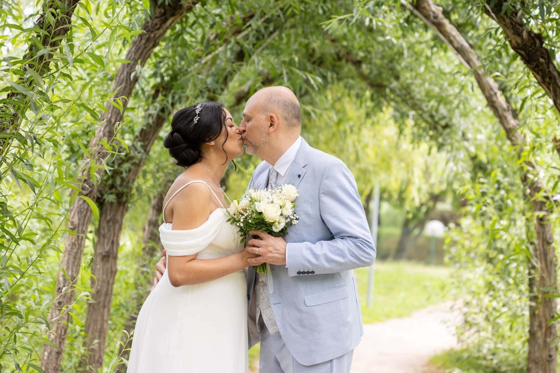 Un couple de mariés s'embrasse dans un jardin verdoyant, la femme porte une robe blanche et tient un bouquet de fleurs, l'homme porte un costume gris clair.