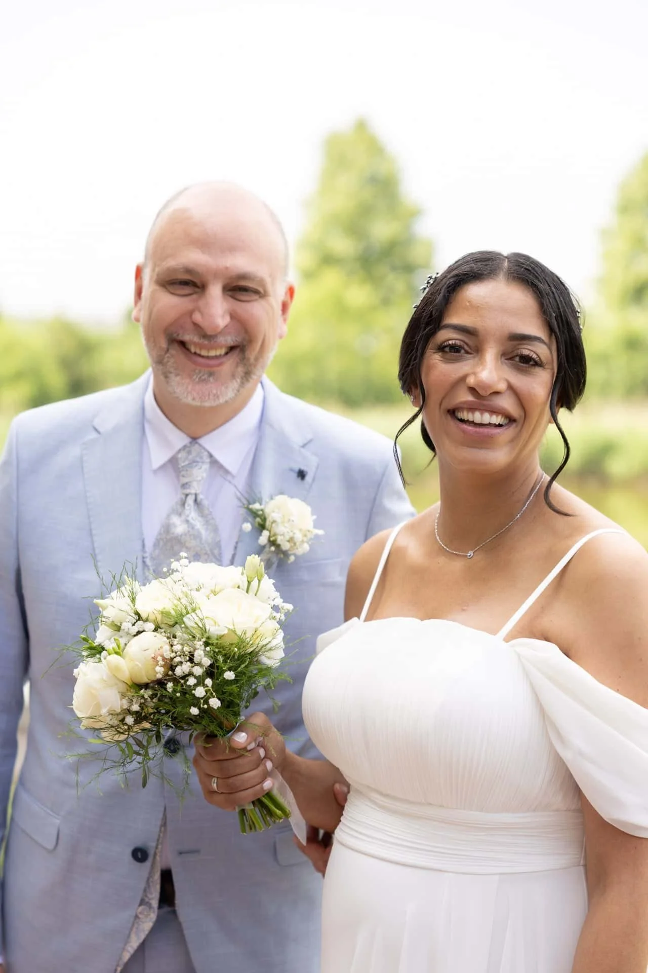 Un couple marié souriant, avec la mariée tenant un bouquet de fleurs blanches, posant à l'extérieur dans un parc verdoyant.