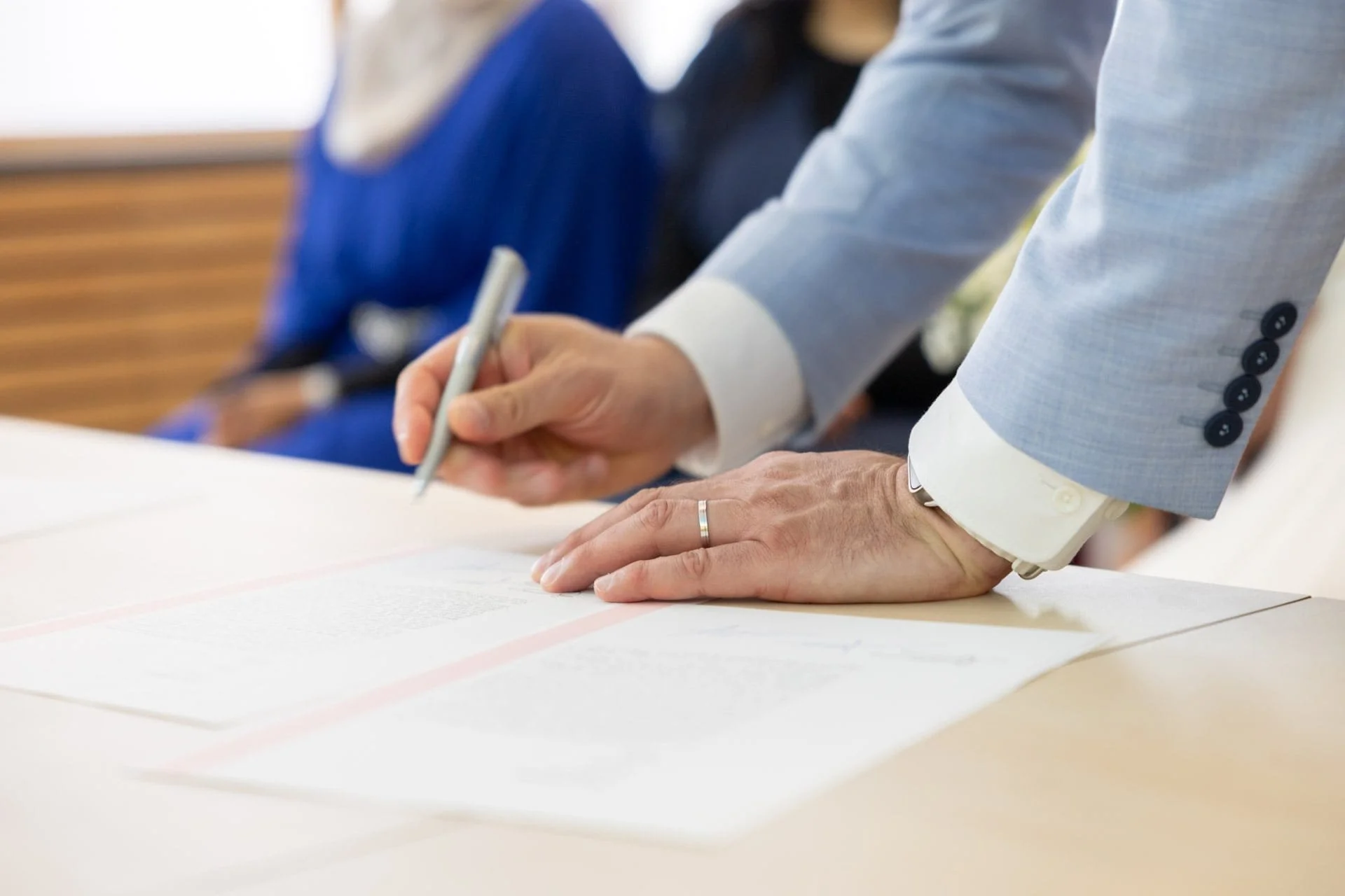 Une personne en costume bleu écrit sur une feuille lors d'une réunion, avec d'autres personnes assises en arrière-plan.